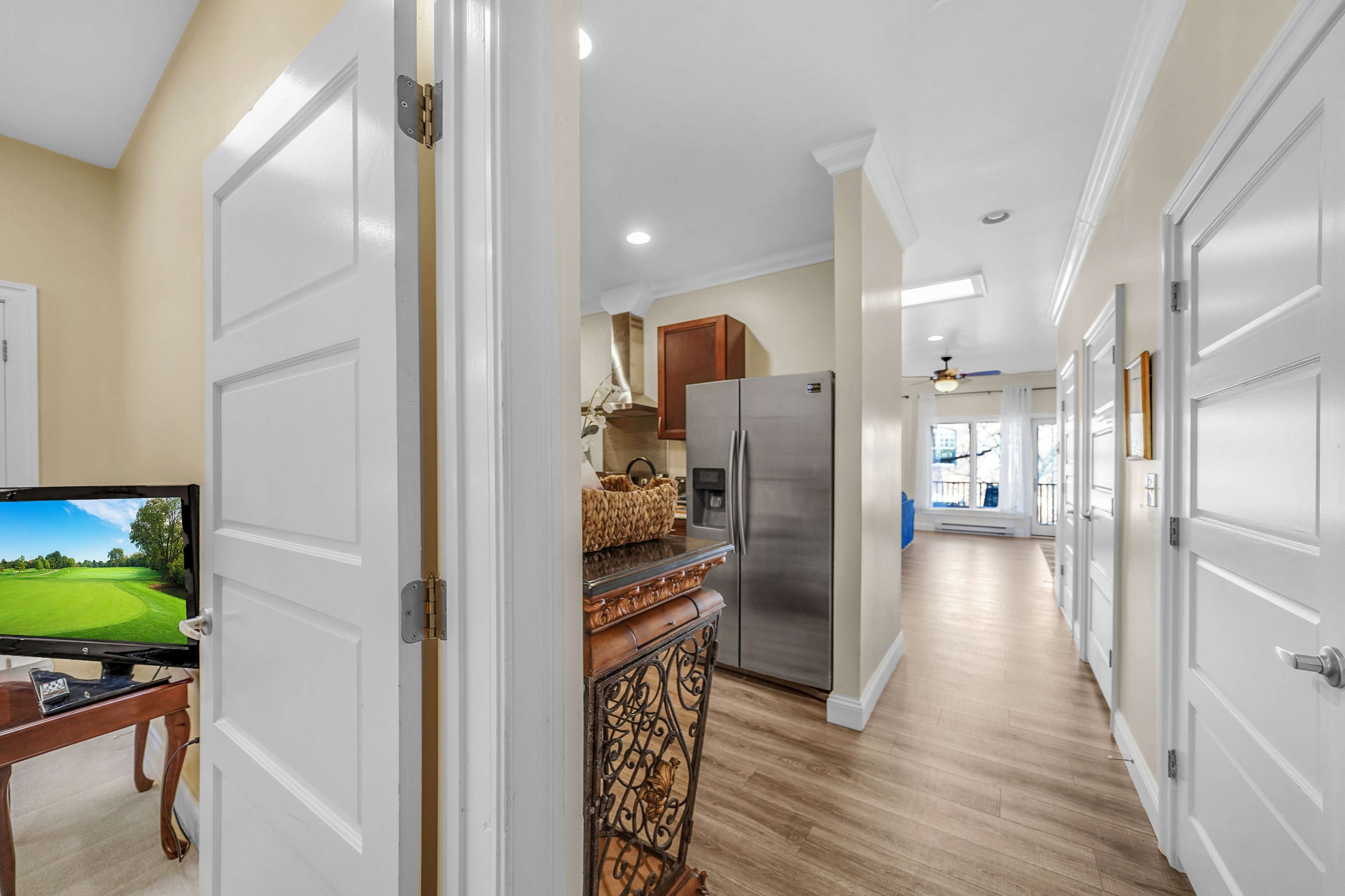 A hallway leading to a kitchen area with a stainless steel refrigerator, a wooden table with a television on it, and a light-filled space at the end.