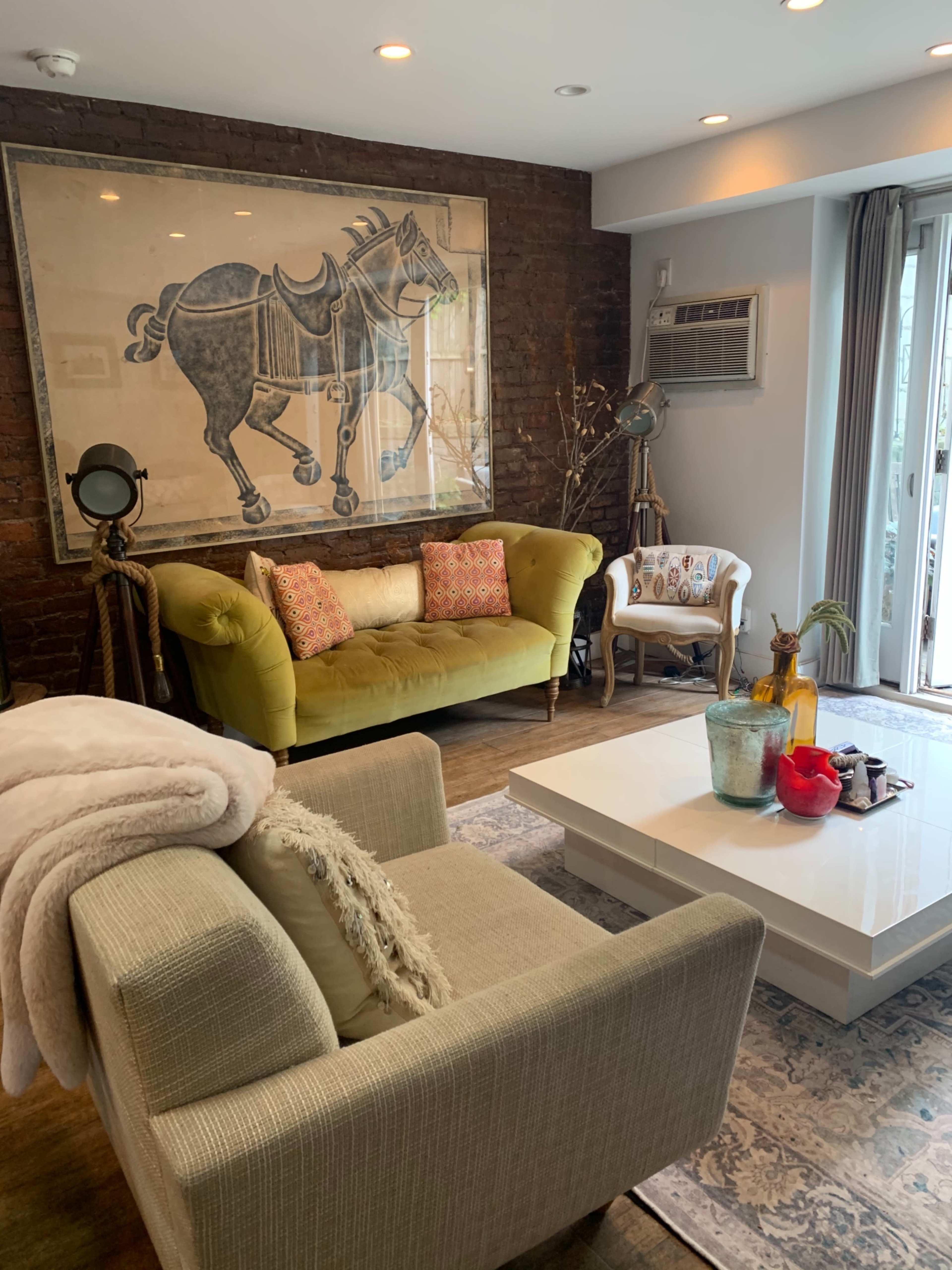 A cozy living room featuring a green sofa, a beige armchair, a large horse painting on the wall, and a white coffee table with decorative items.