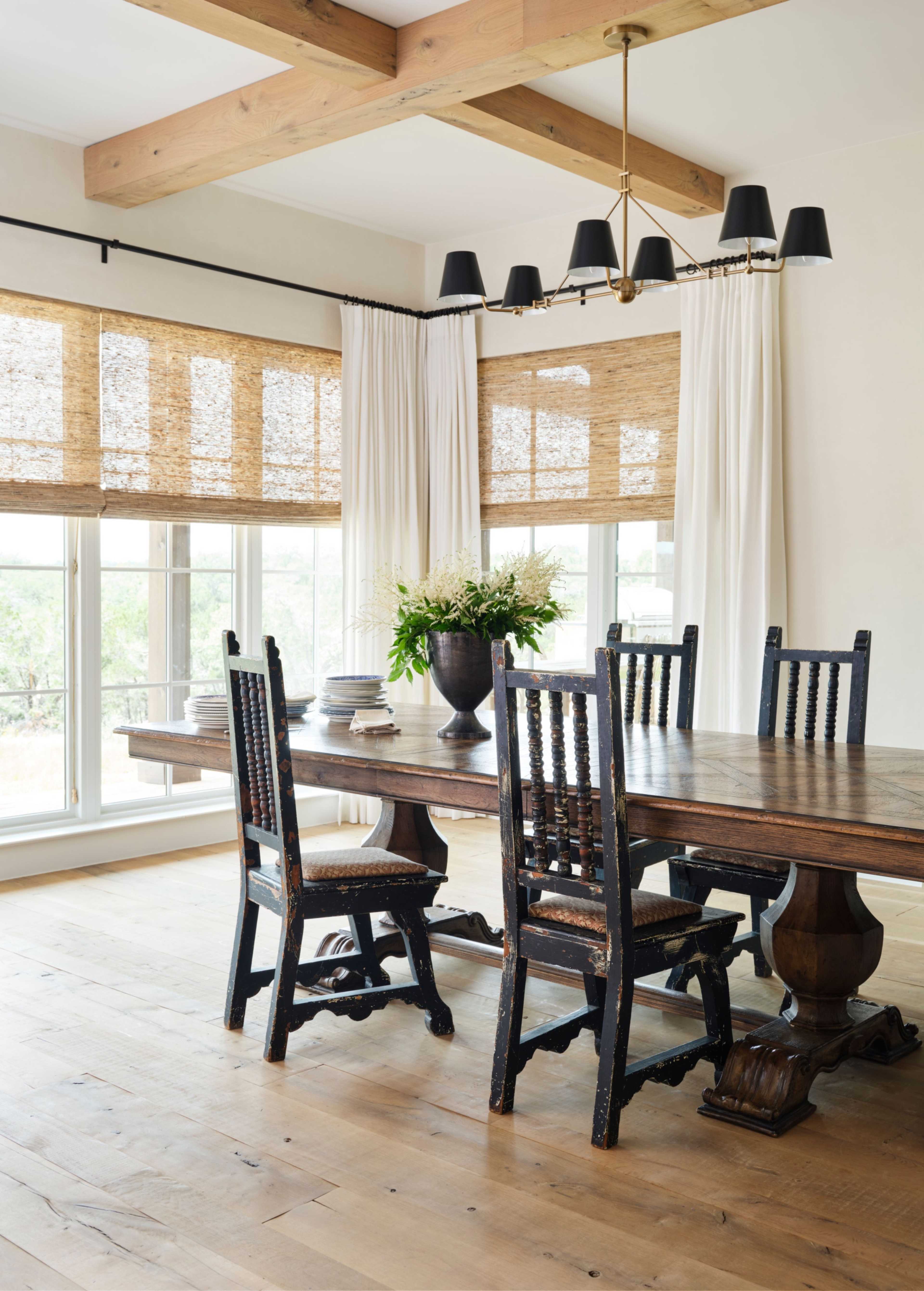 A dining room features a wooden table surrounded by black chairs, large windows with natural light, and light-colored curtains.