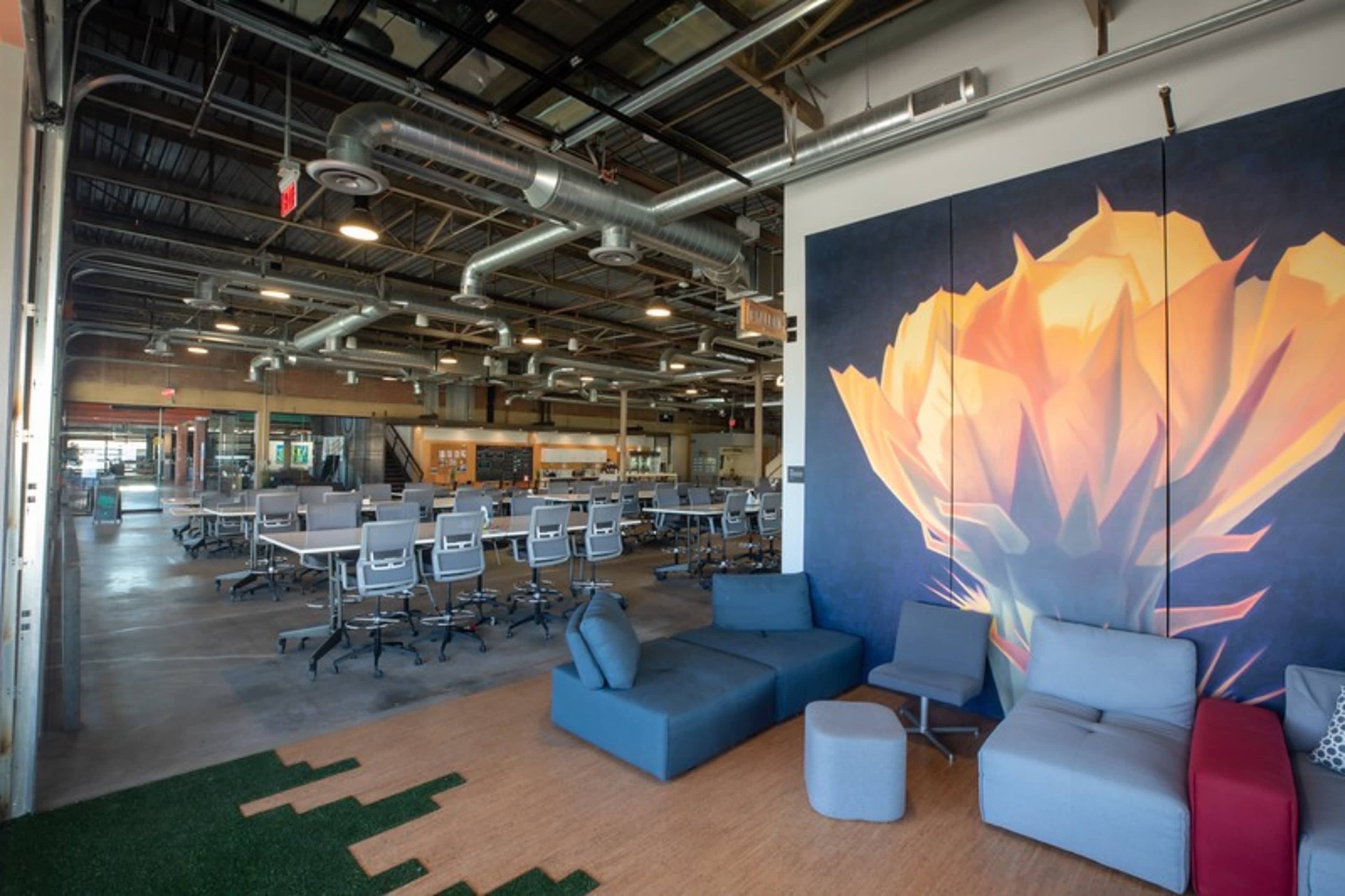 A modern open office space with a large flower mural, featuring a communal workspace with multiple desks and ergonomic chairs, along with a seating area.