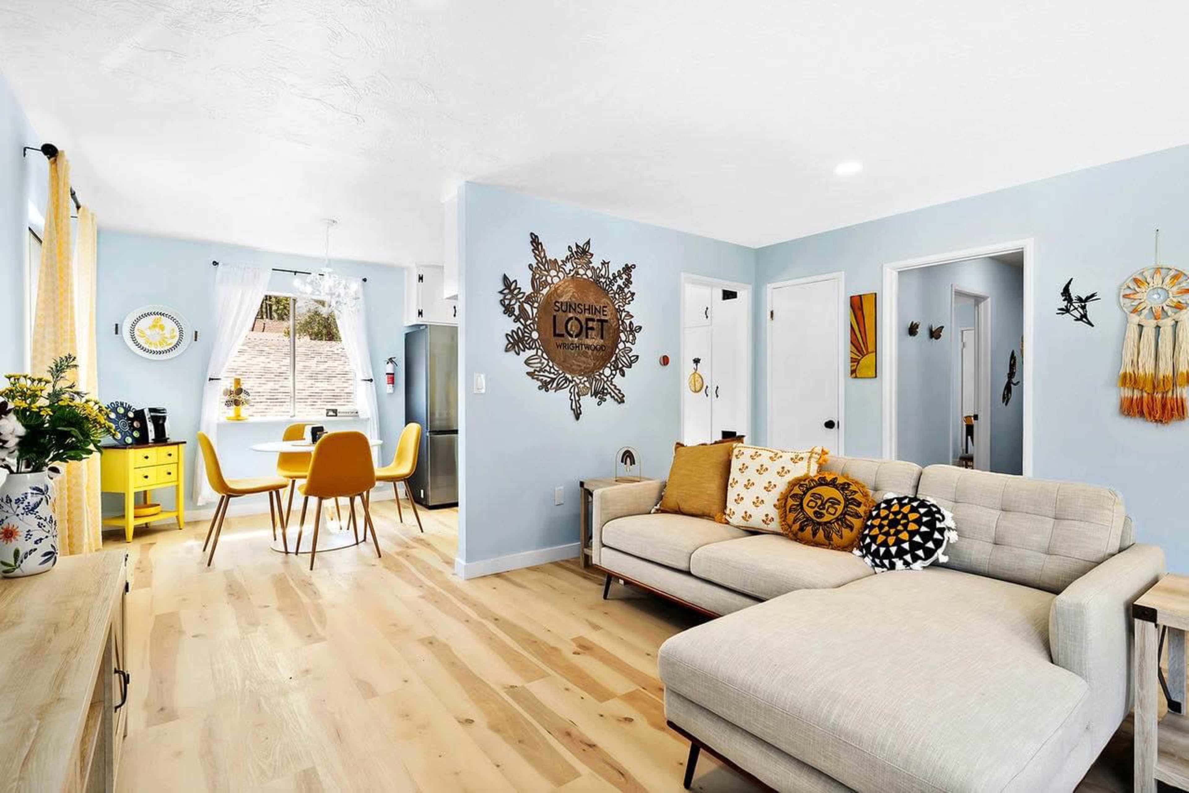 A bright living area with a sectional sofa, a yellow dining table with chairs, and decorative wall elements amidst light blue walls and wooden flooring.