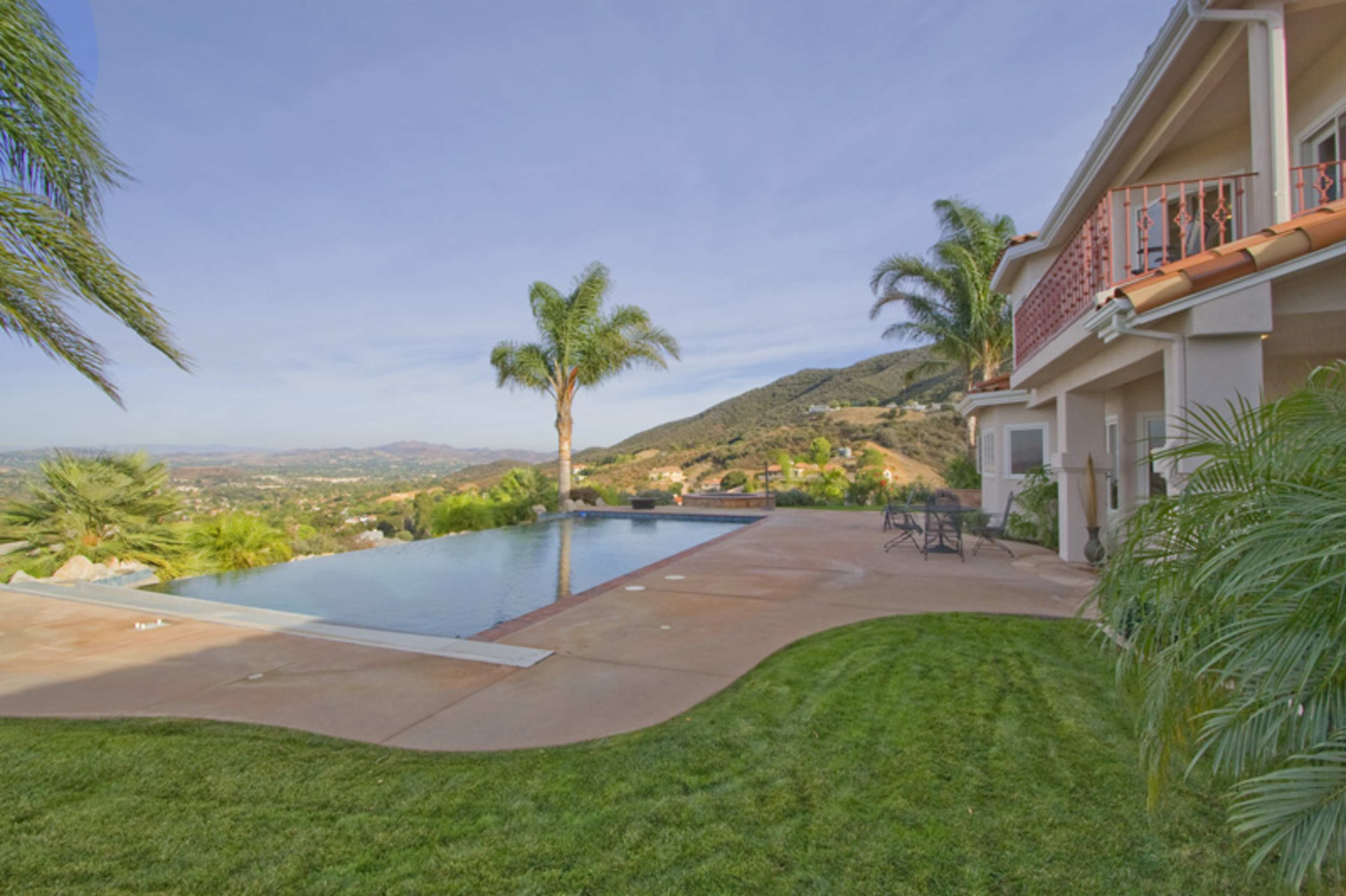 A modern house sits beside an infinity pool overlooking a mountainous landscape with palm trees and a lawn.