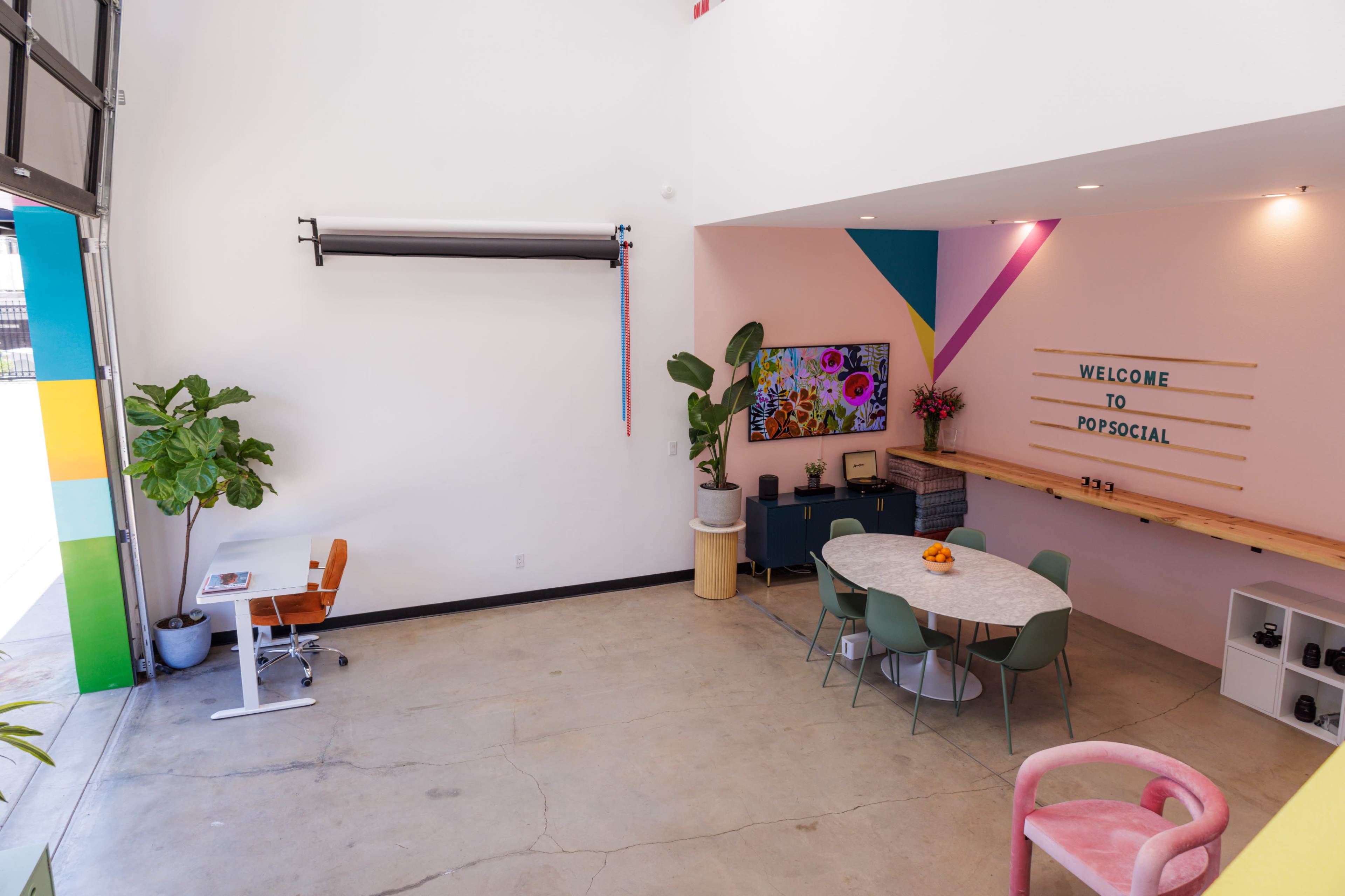 A brightly lit interior features a round marble table surrounded by chairs, a large television on the wall displaying colorful artwork, and a workspace in one corner.