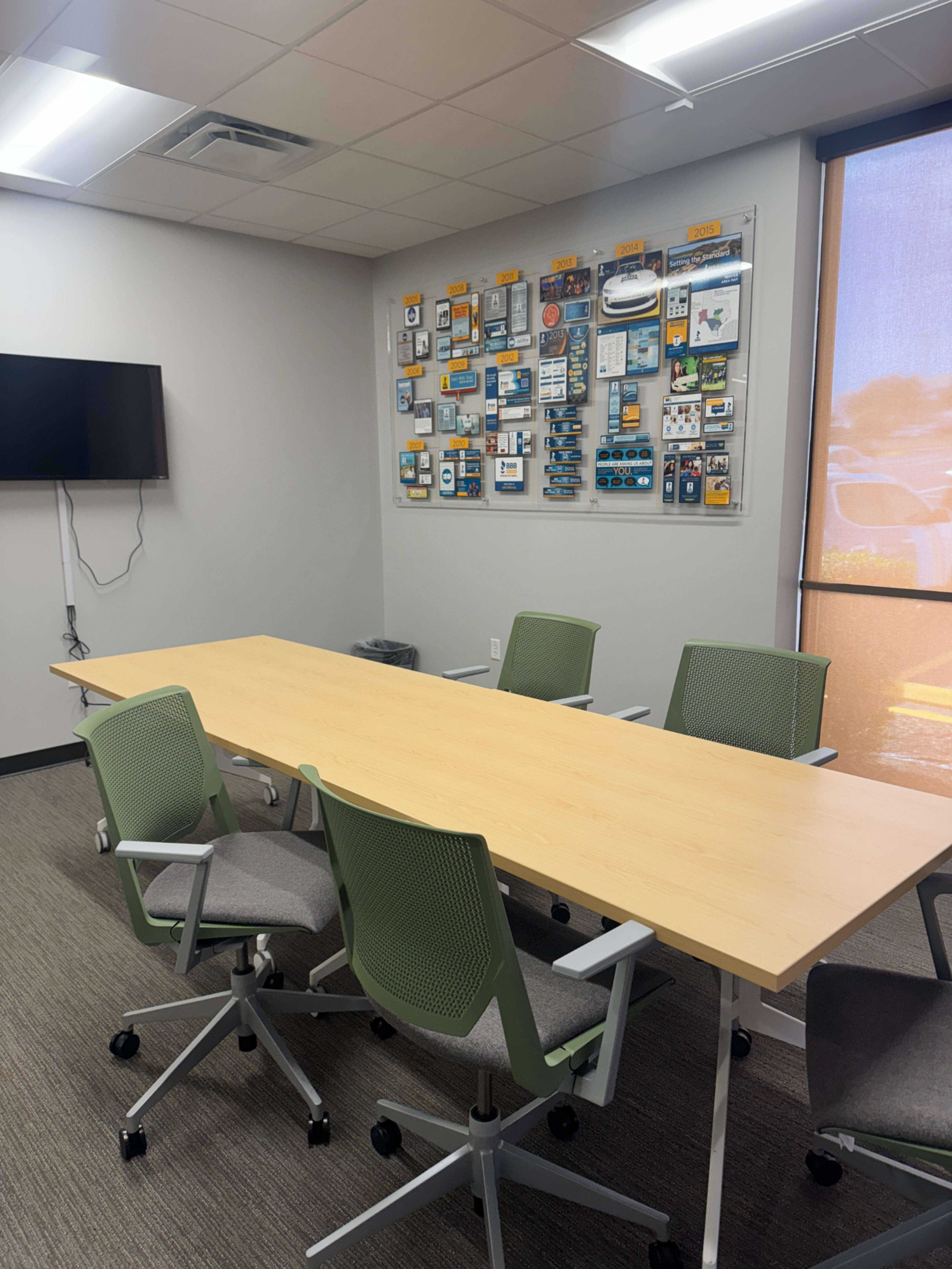 A conference room features a long wooden table surrounded by green chairs, with a wall displaying various technology items and a TV mounted nearby.