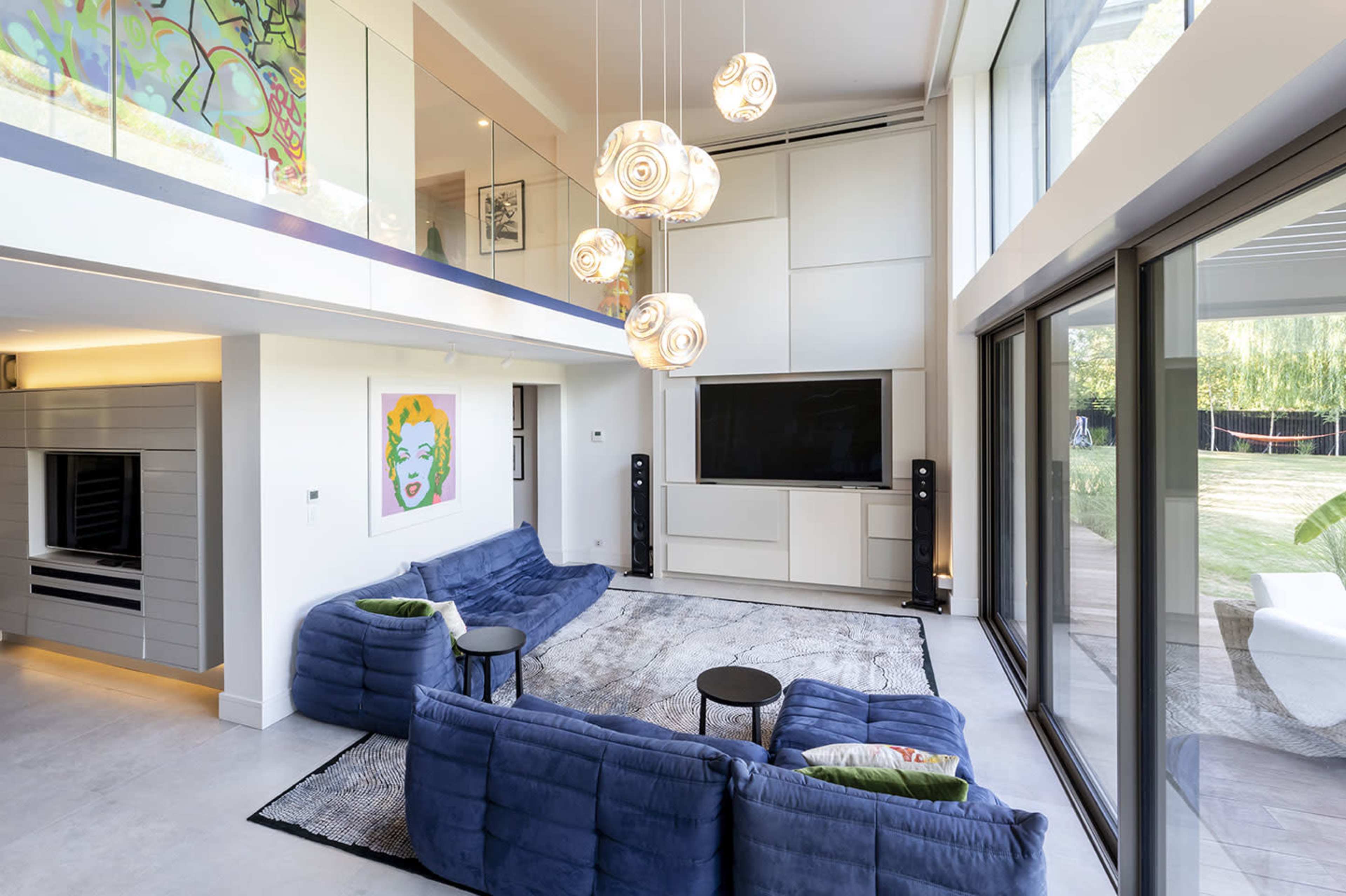 A modern living room with blue sofas, pendant lights, and large glass doors overlooking a garden.