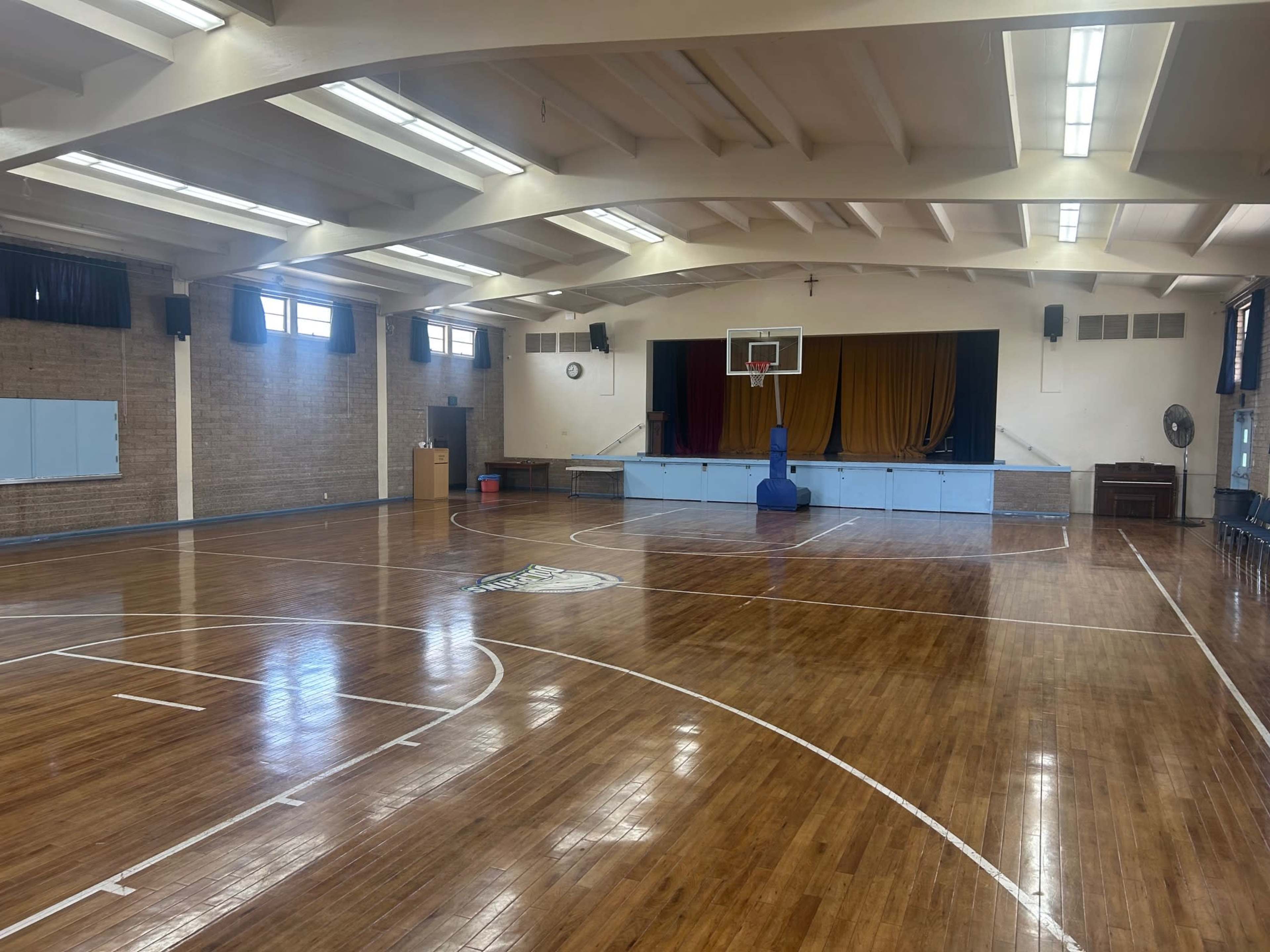The image shows a spacious indoor basketball court with polished wooden floors, a basketball hoop, and an adjacent stage area.