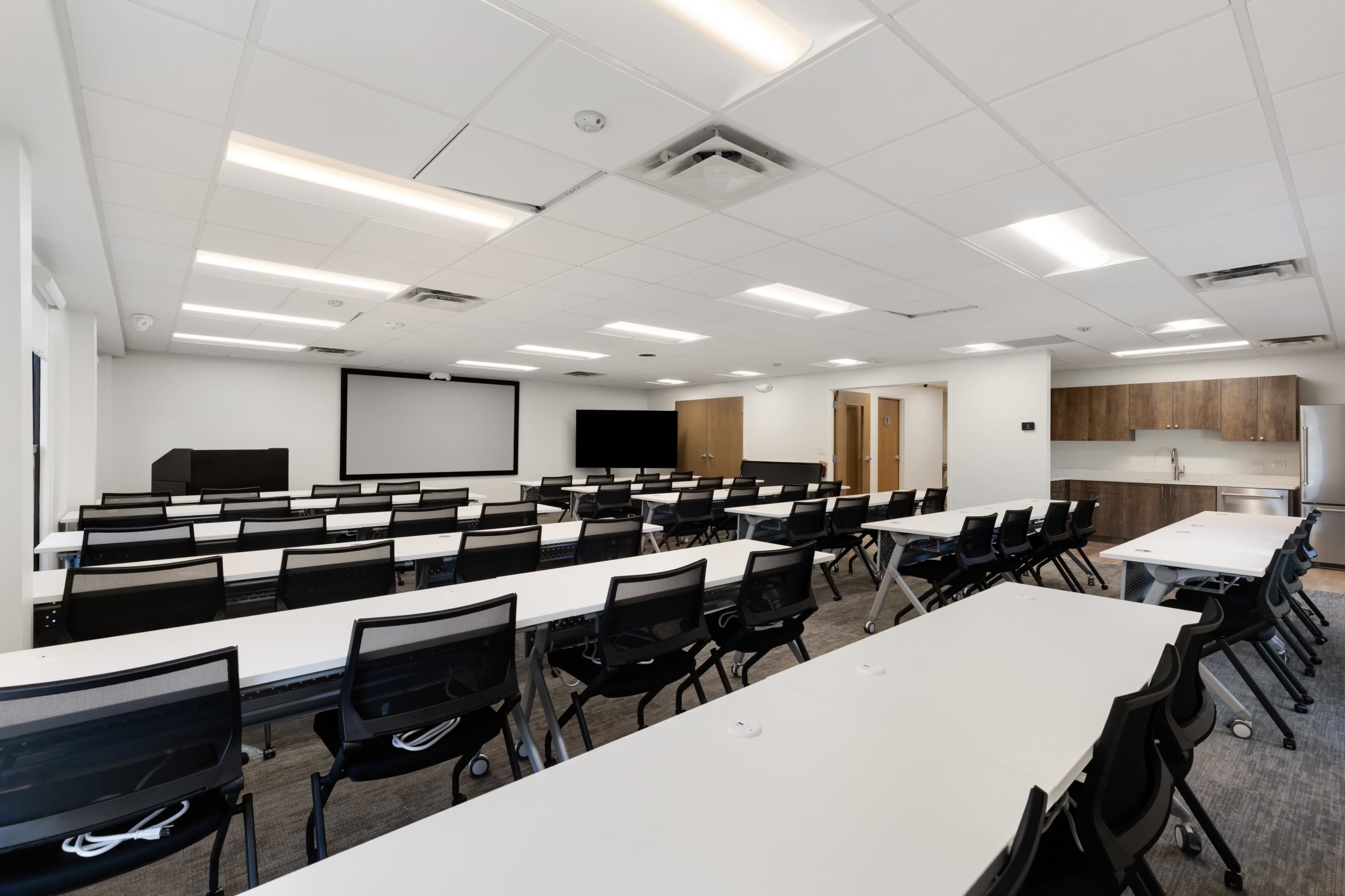 The image shows a modern conference room featuring rows of tables and chairs, two screens on the front wall, and a kitchenette in the corner.