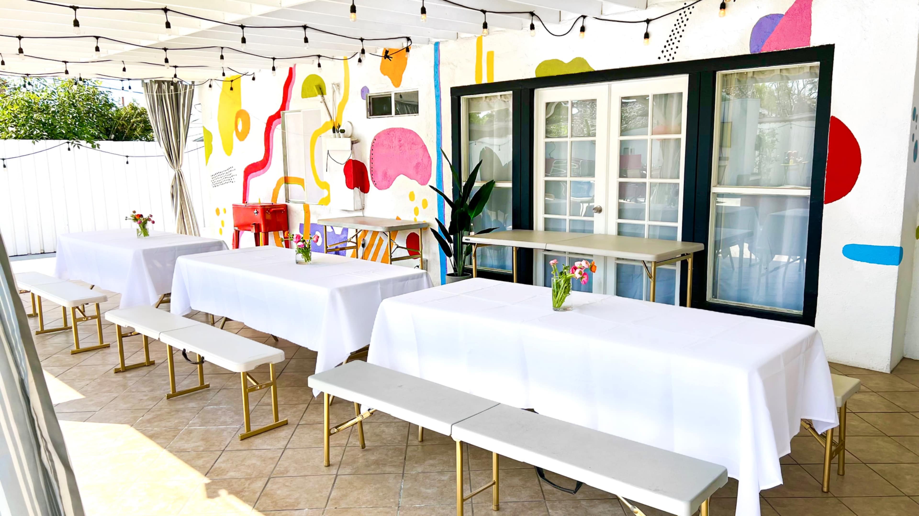 A patio area is set with white tablecloths on multiple tables, surrounded by colorful wall art and string lights.