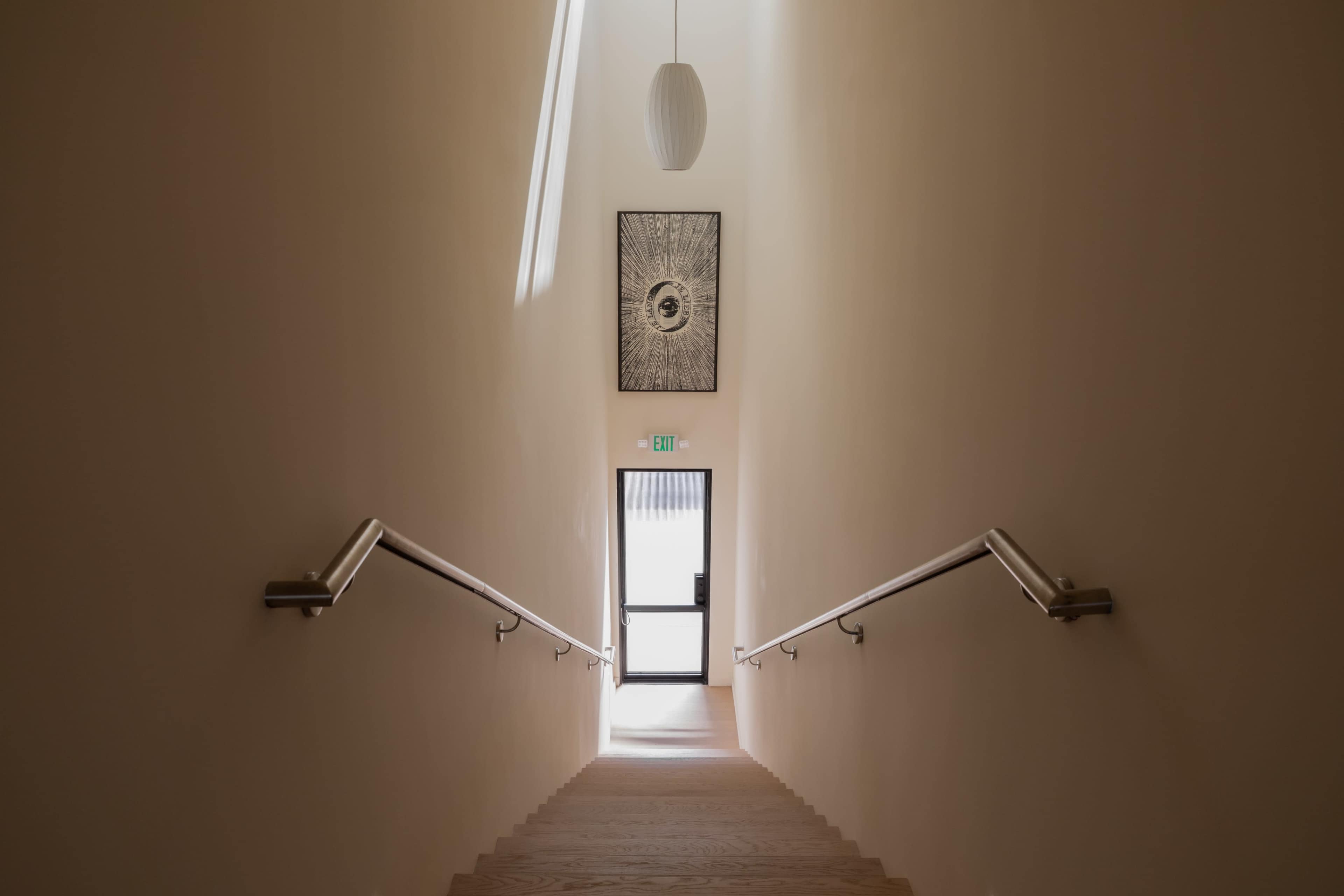 A well-lit staircase leads down to a door at the bottom, illuminated by a hanging light and adorned with a piece of art on the wall.