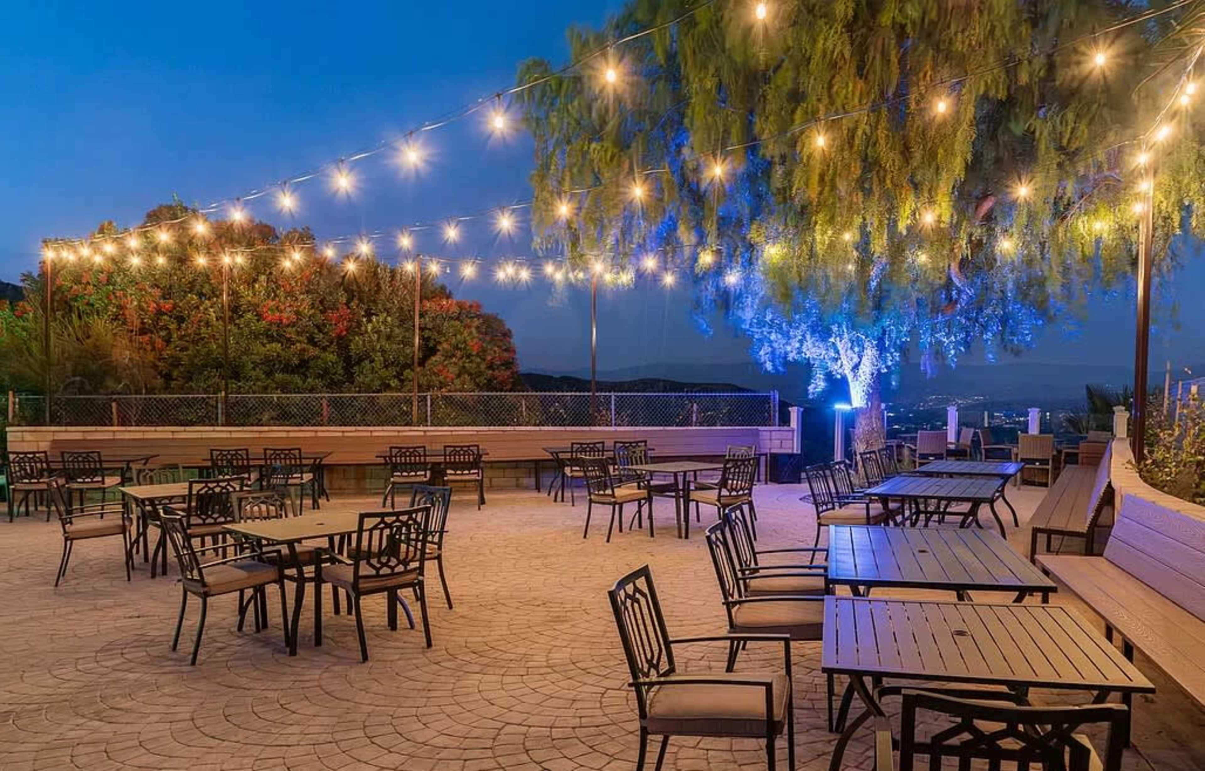 An outdoor dining area features numerous tables and chairs arranged beneath string lights and greenery.
