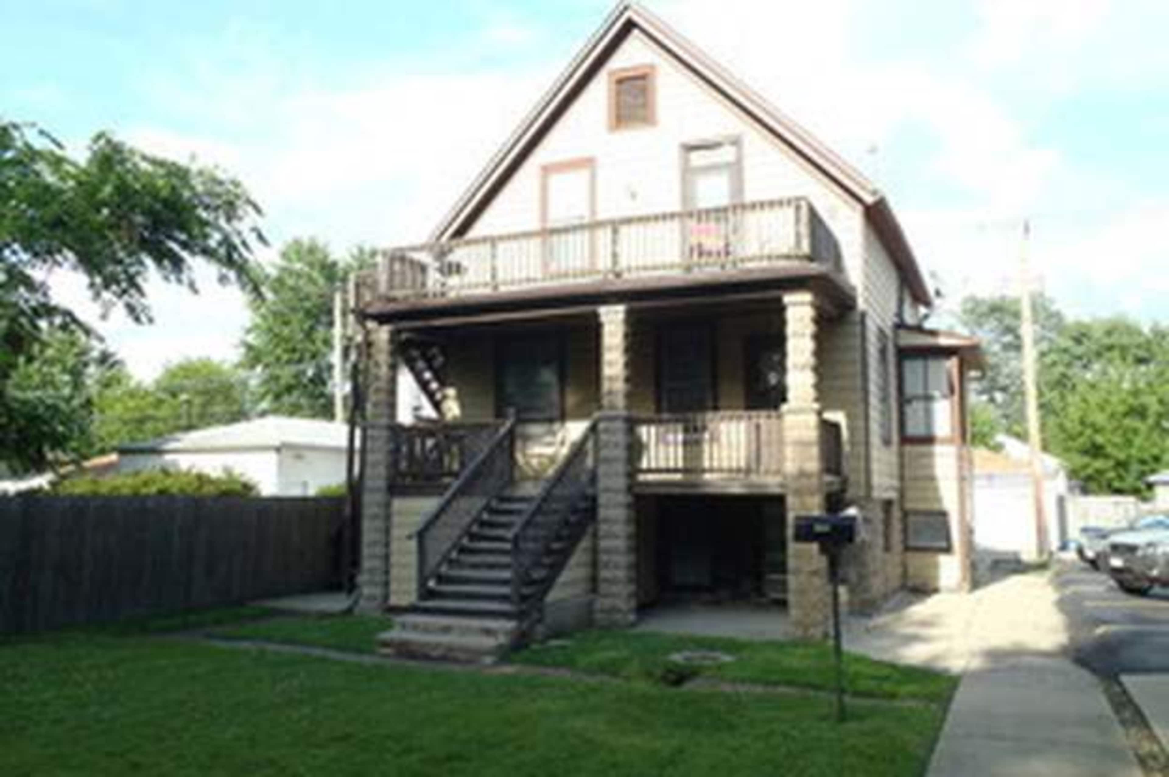 A two-story house with a front porch, wooden railings, and a staircase leading to the entrance is surrounded by grass and a concrete pathway.