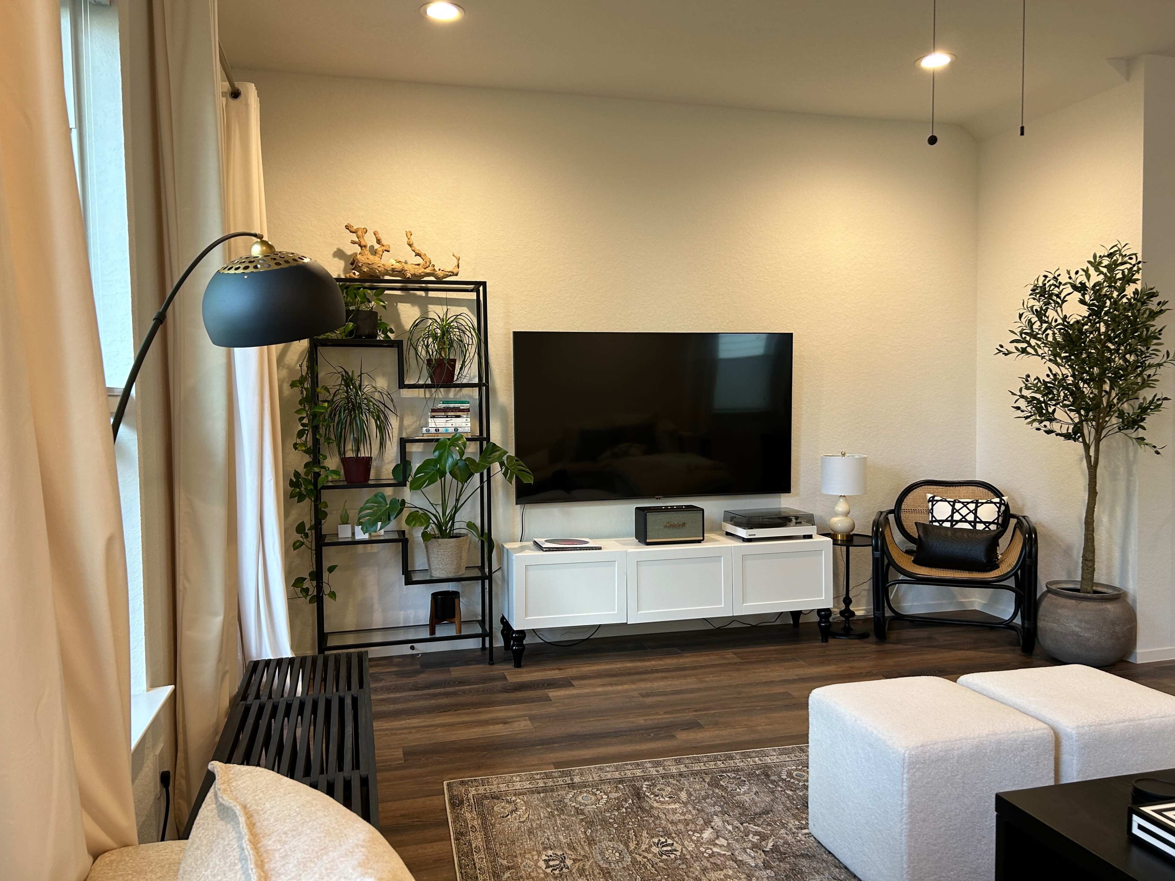 A modern living room features a large TV mounted on a beige wall, surrounded by plants, a bookshelf, and a white cabinet with a sleek black lamp and a wicker chair.