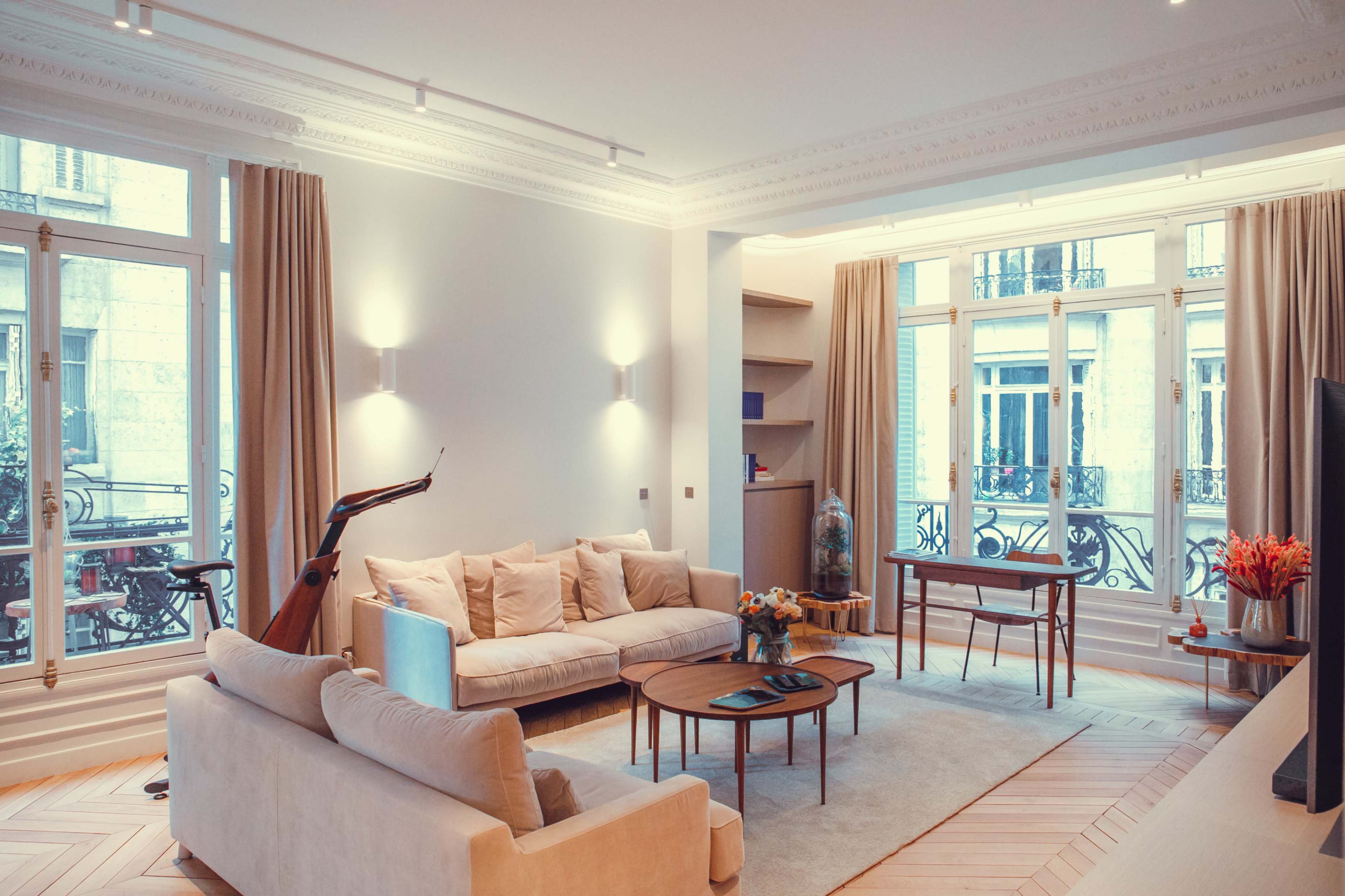 The image shows a spacious living room with large windows, a beige sofa, a coffee table, and a small work desk.