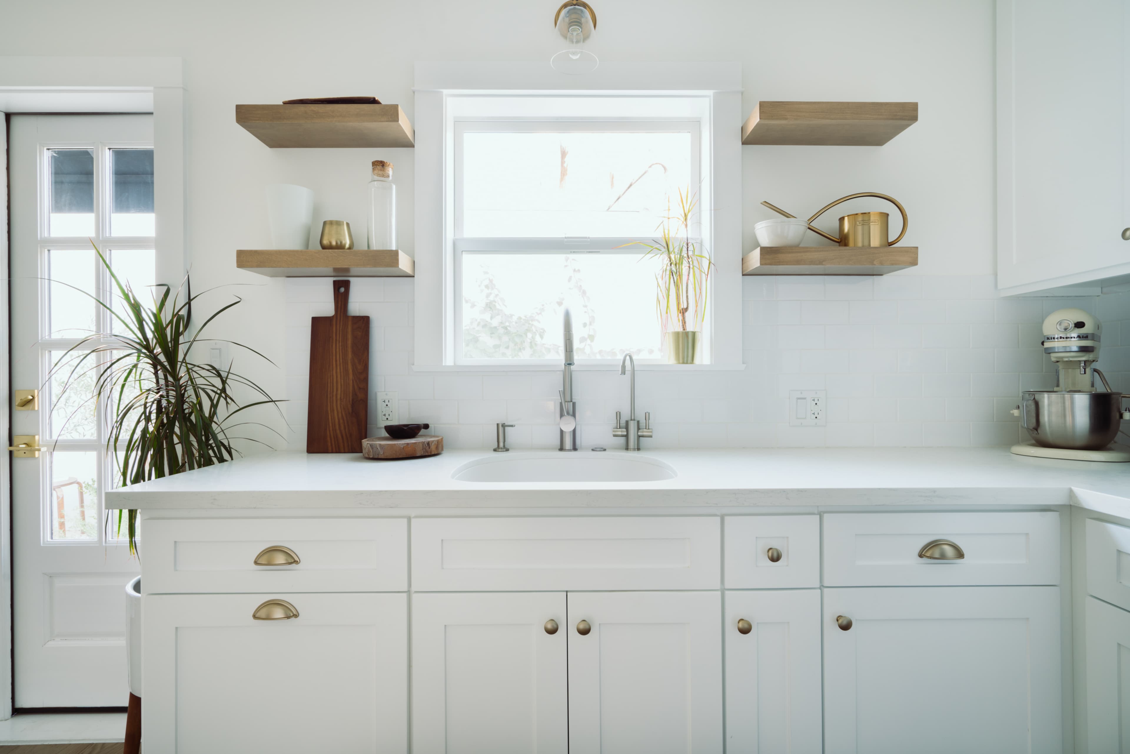 The image shows a bright kitchen featuring a white sink, white cabinetry, wooden shelves, and a window with plants visible outside.