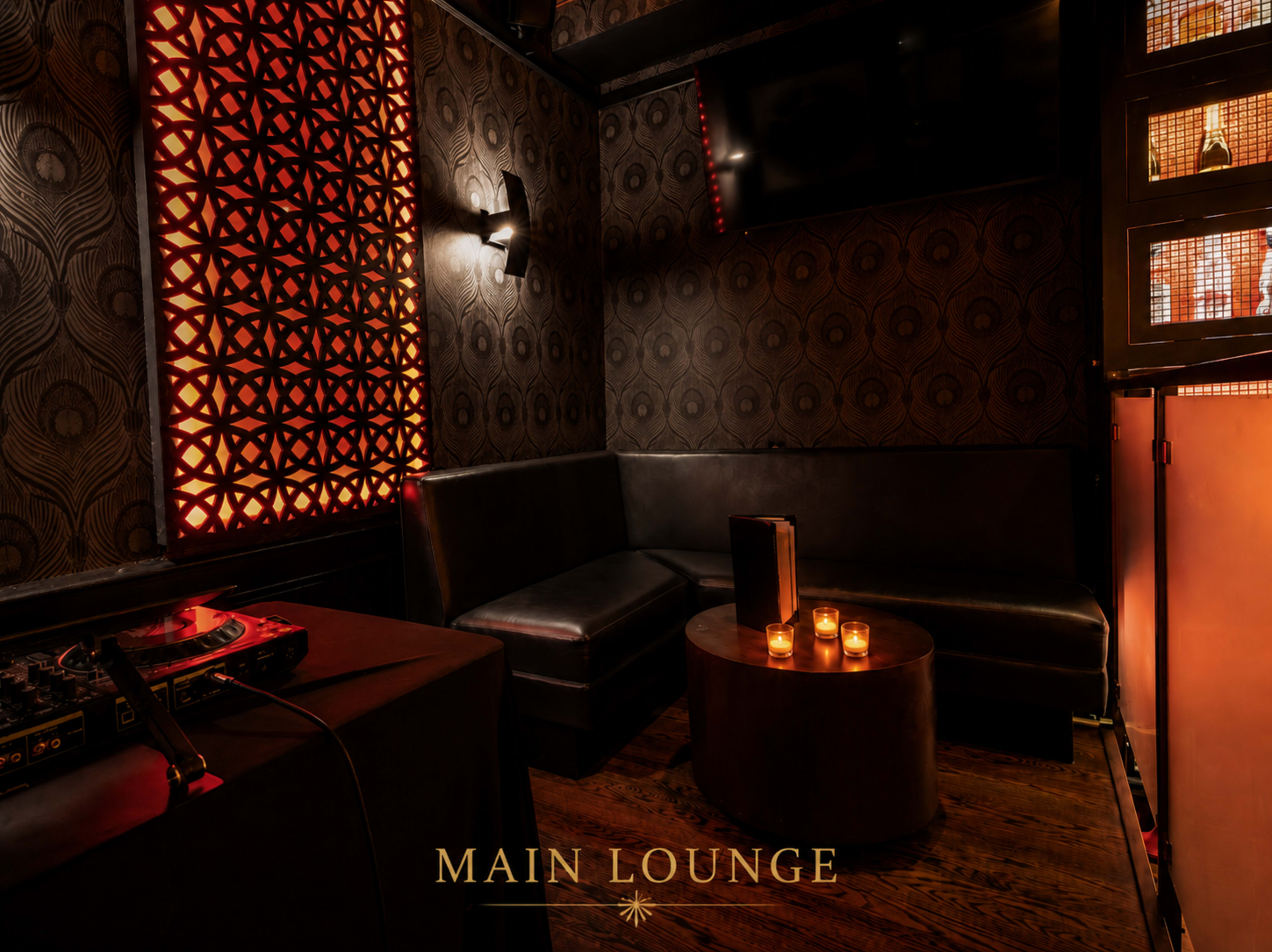 The image shows a dimly lit lounge area featuring a dark upholstered seating arrangement, a round table with candles, and a DJ setup in the corner.