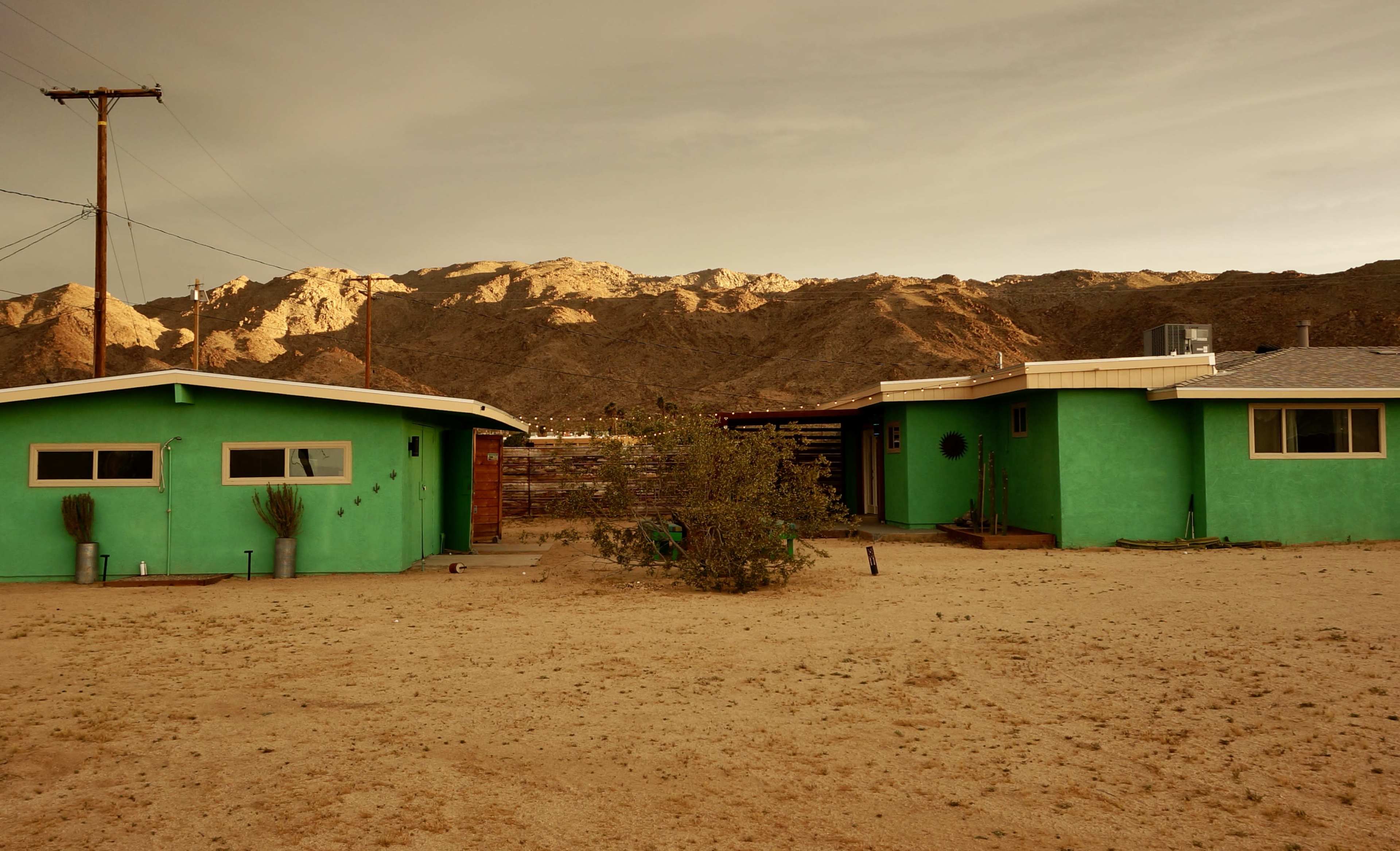 Two green houses are situated in a sandy landscape with mountains in the background under a cloudy sky.