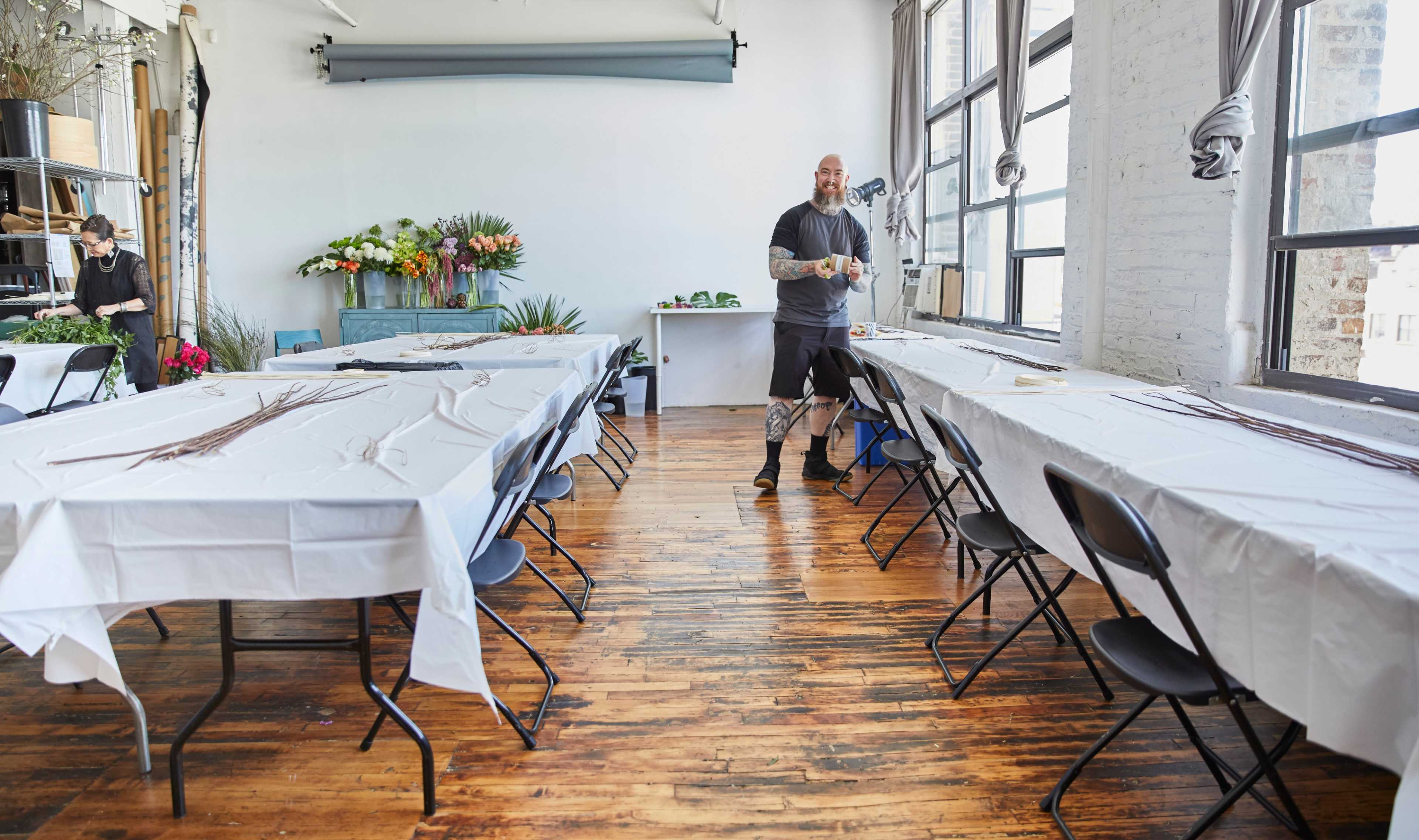 A man prepares materials in a spacious workshop filled with tables covered in white cloth, with flower arrangements visible in the background.