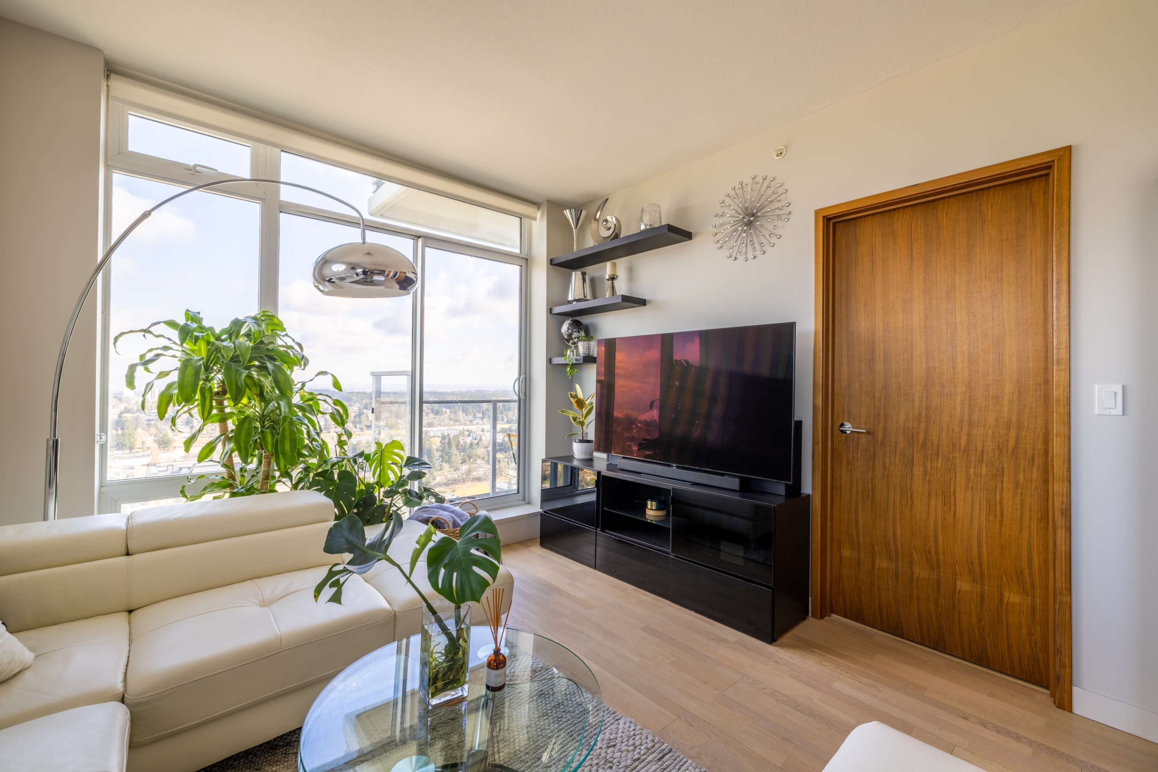 A modern living room with a white leather sofa, a glass coffee table, a large TV on a black stand, a potted plant, and a wooden door leading outside.
