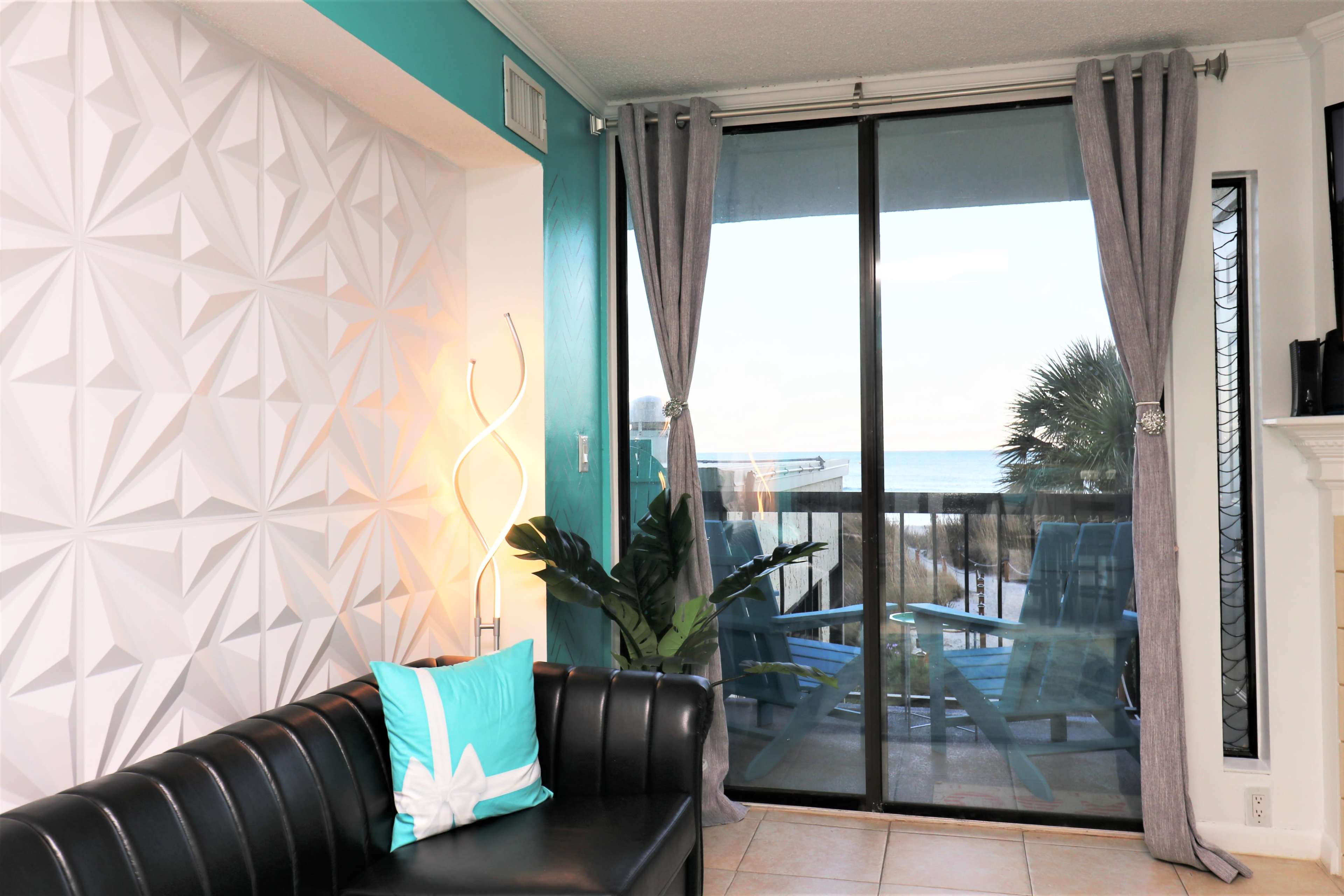 A modern living room with a black couch, a decorative light, and a large glass door that opens to a balcony with an ocean view.