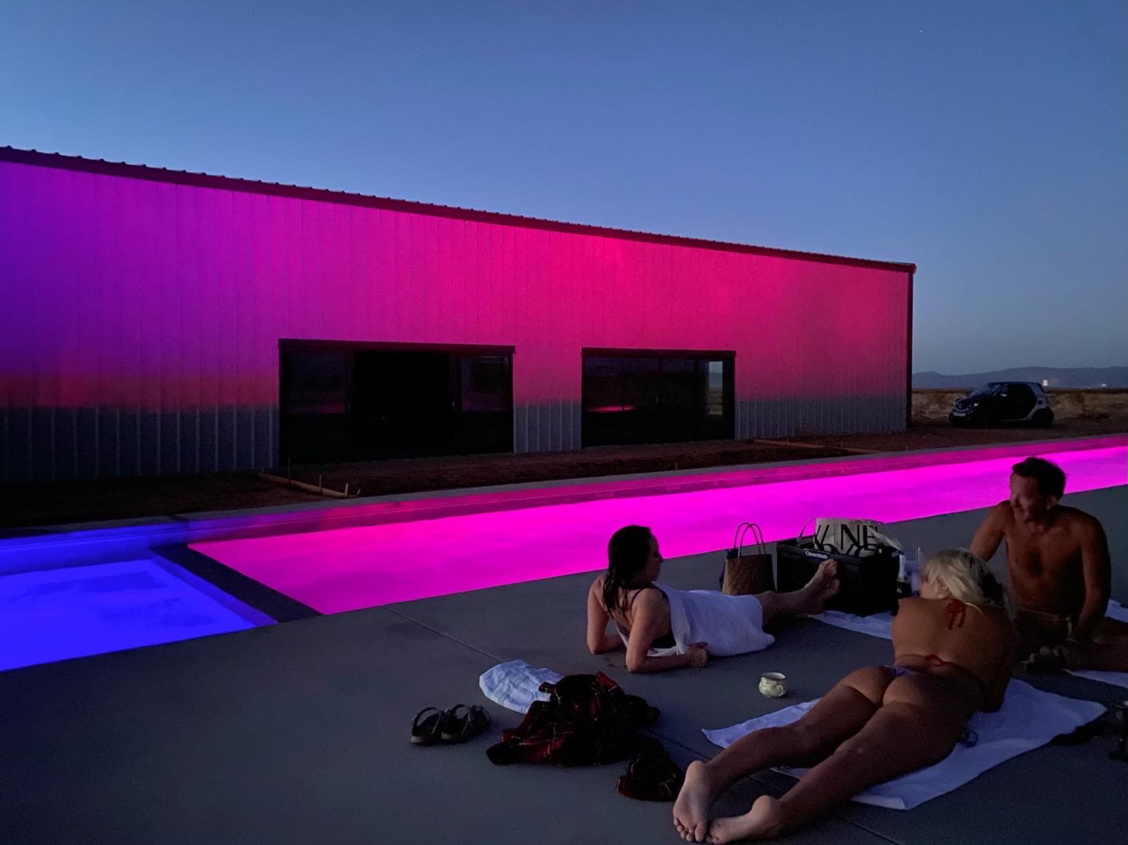 Three people lounge by a brightly illuminated swimming pool at dusk, with a modern building reflecting pink and purple lights in the background.