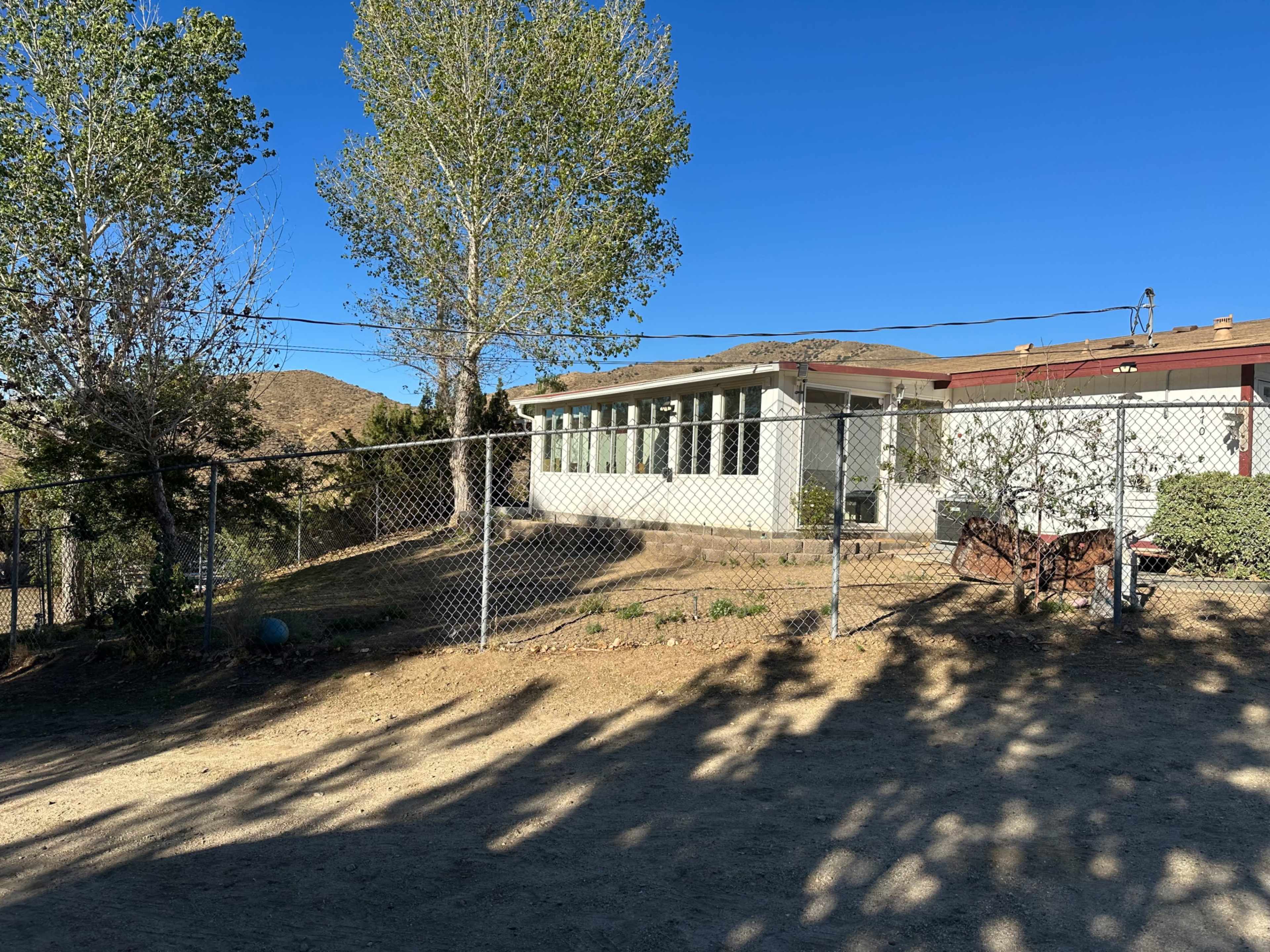 A fenced yard with a house featuring large windows and surrounding trees against a clear blue sky.