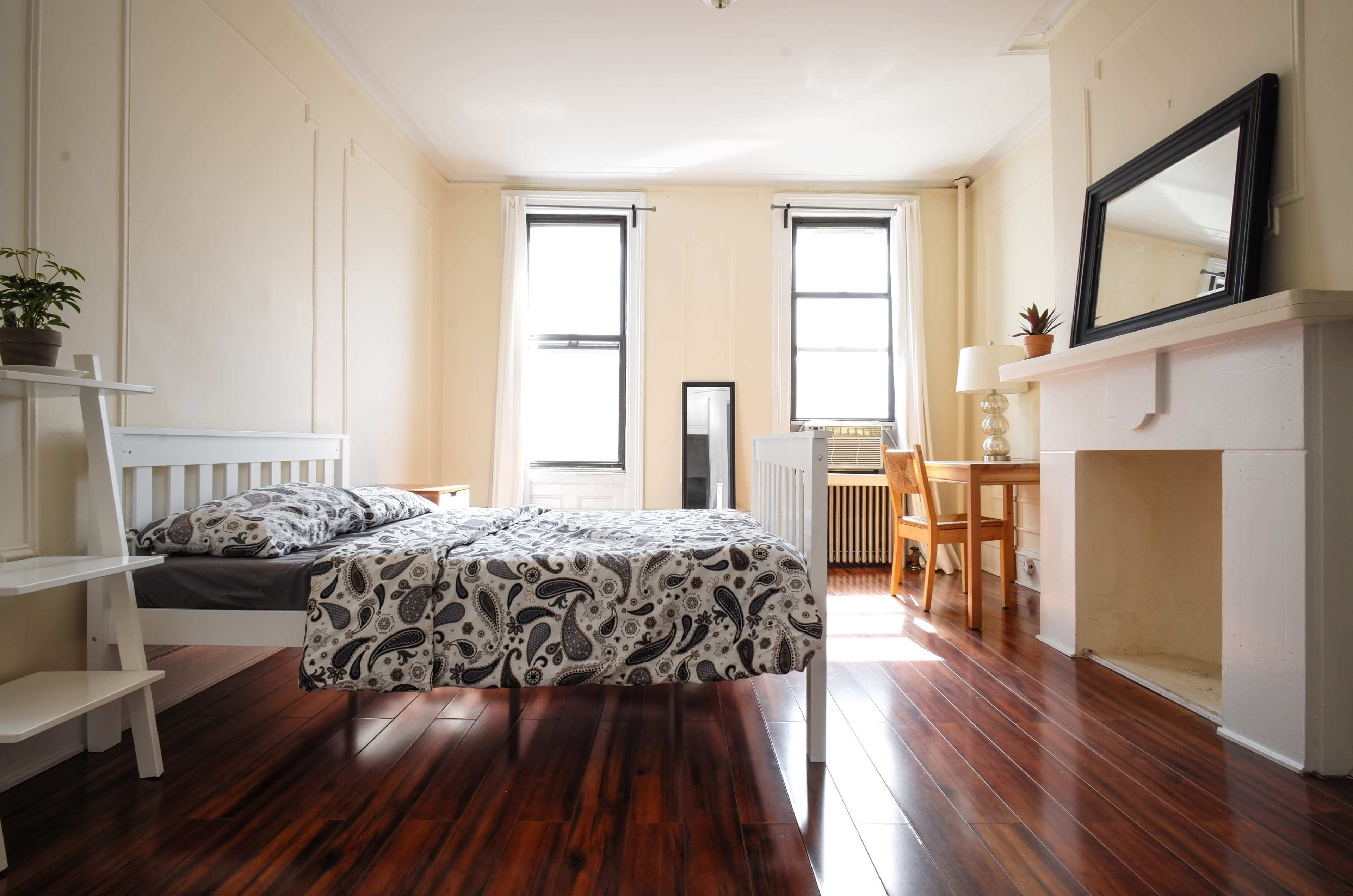 The room features a bed with a patterned comforter, a wooden chair and table, a fireplace, and large windows allowing natural light to enter.