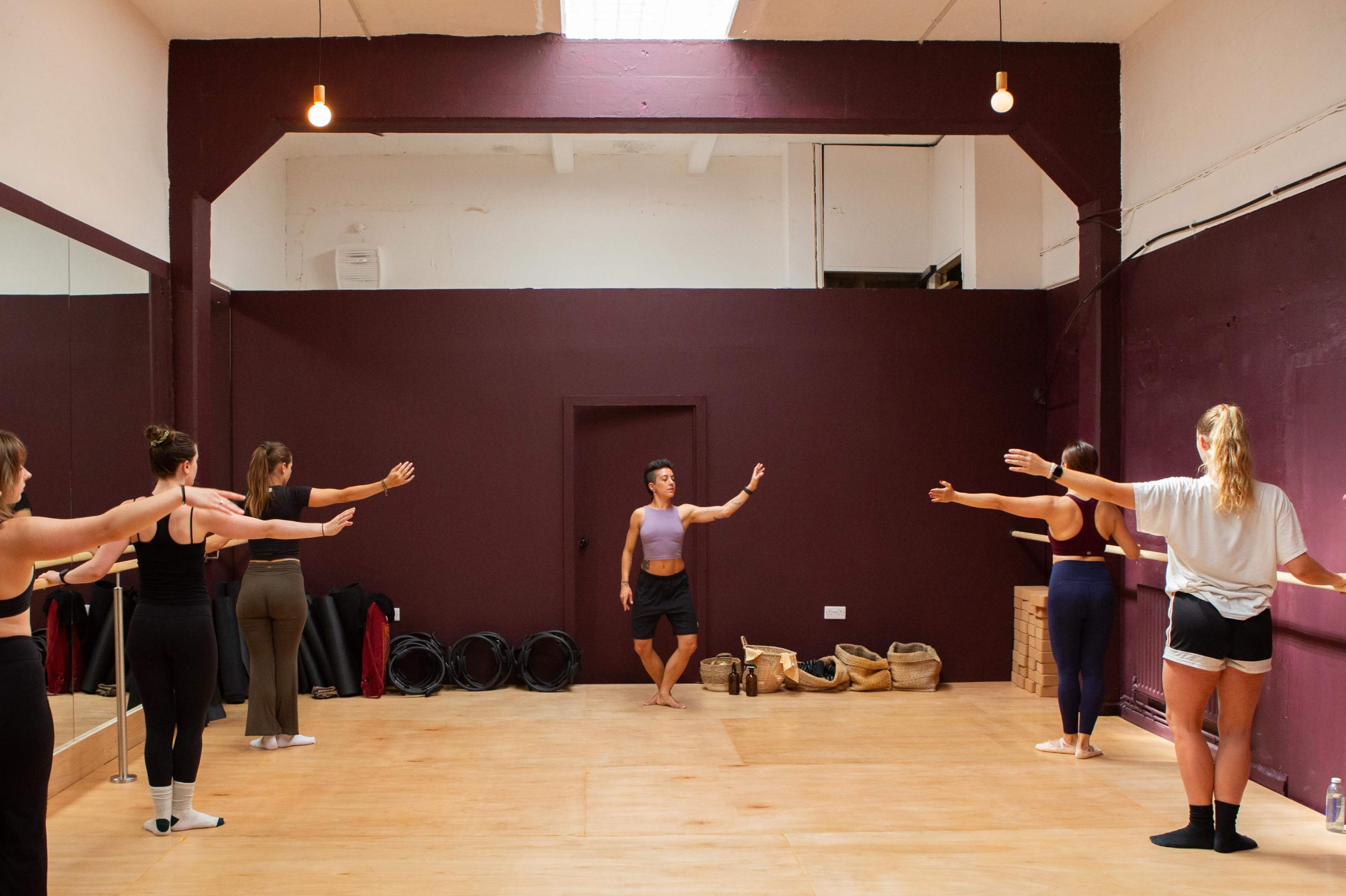 A dance instructor demonstrates a pose to a group of students in a mirrored studio with wooden floors and a purple accent wall.