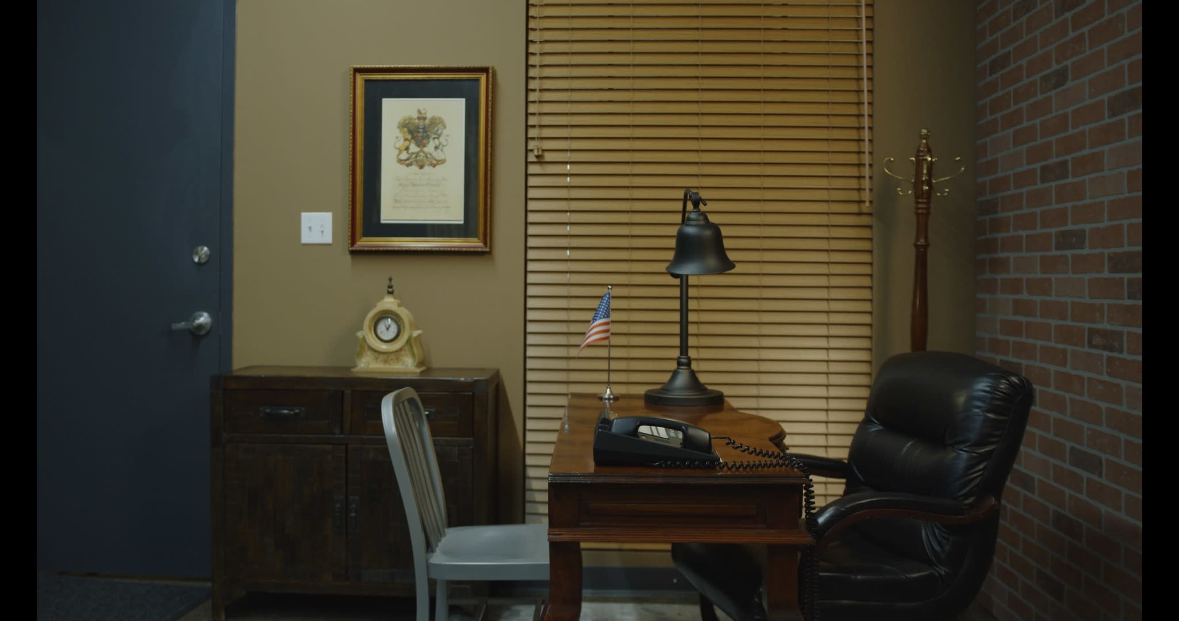 A desk with a lamp and a rotary phone is positioned in a corner of an office, accompanied by a chair and a small American flag on the desk, with a framed document on the wall above it.