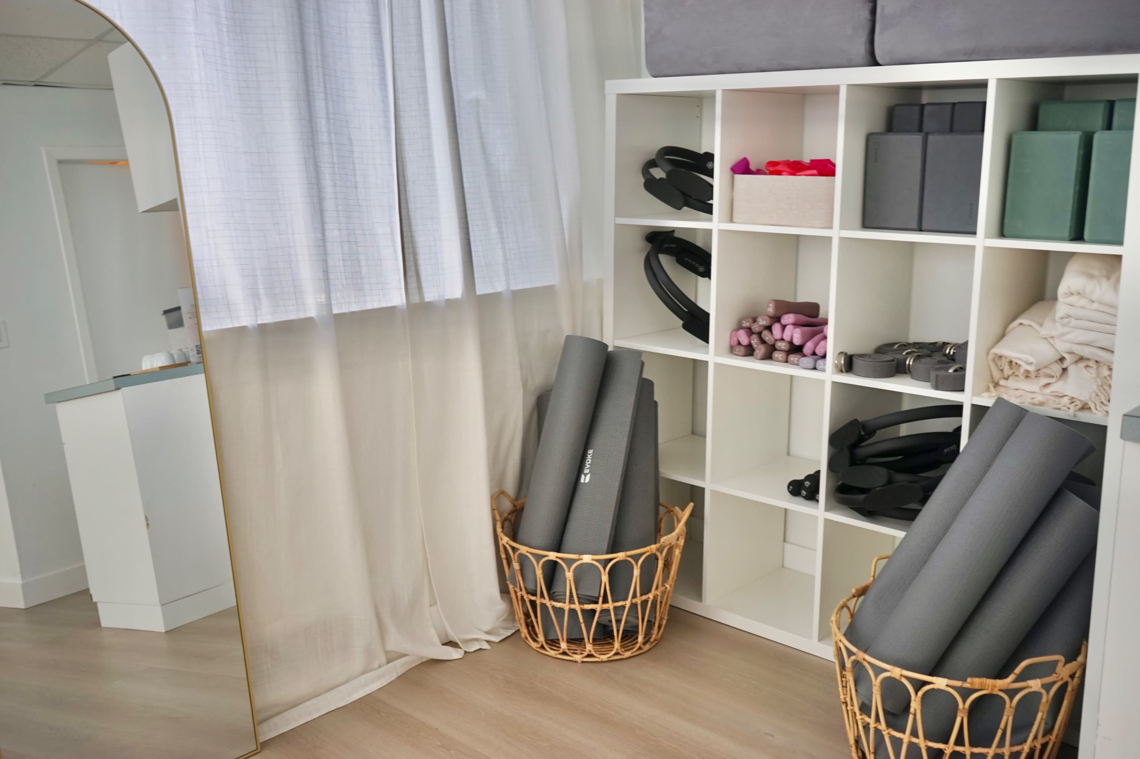 The image shows a bright room with a shelving unit containing neatly arranged fitness equipment, yoga mats, and towels beside two woven baskets holding rolled exercise mats.