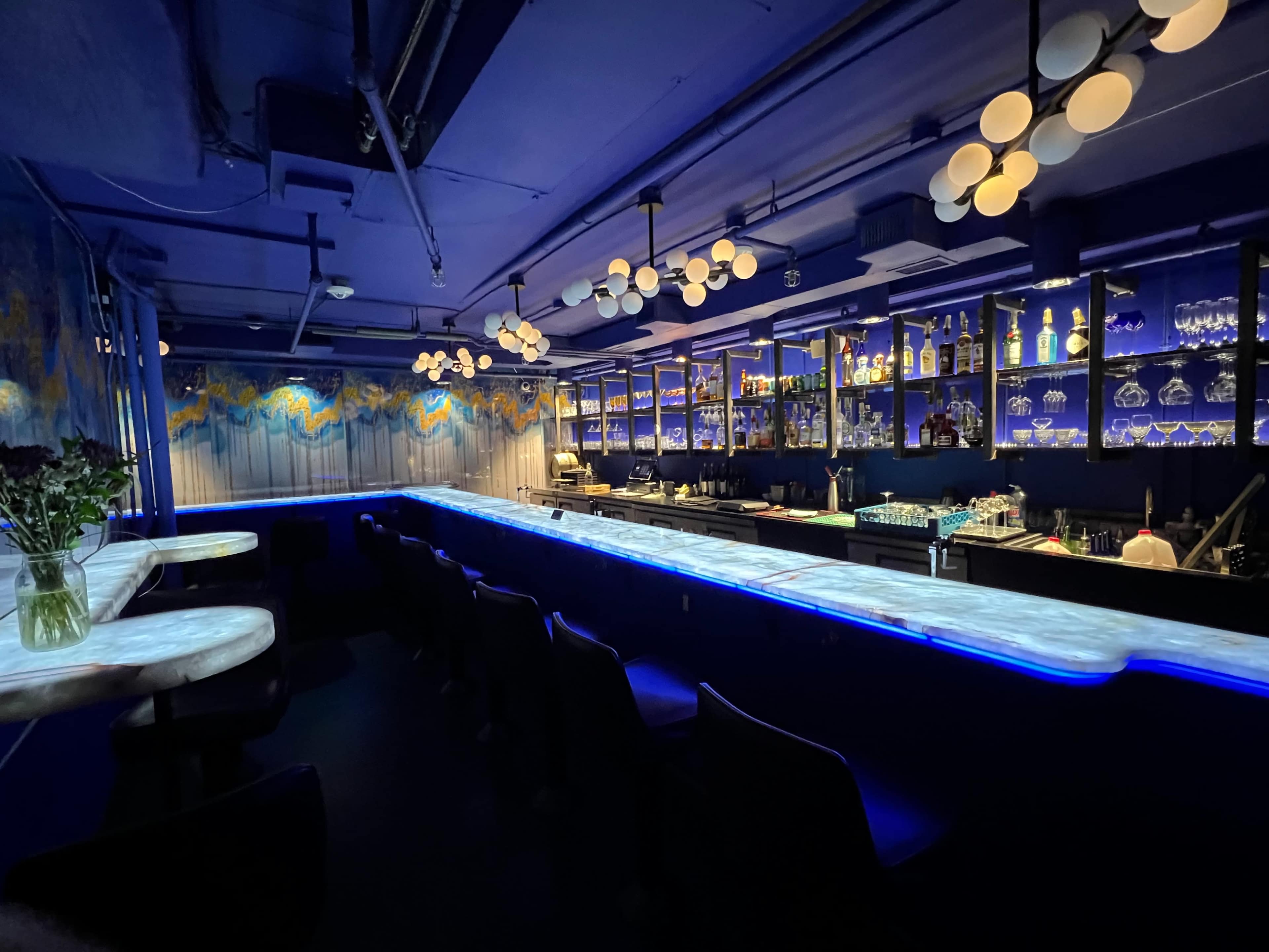 The image shows a modern bar interior with a glowing blue countertop, shelves filled with glassware and bottles, and a deep blue color scheme.