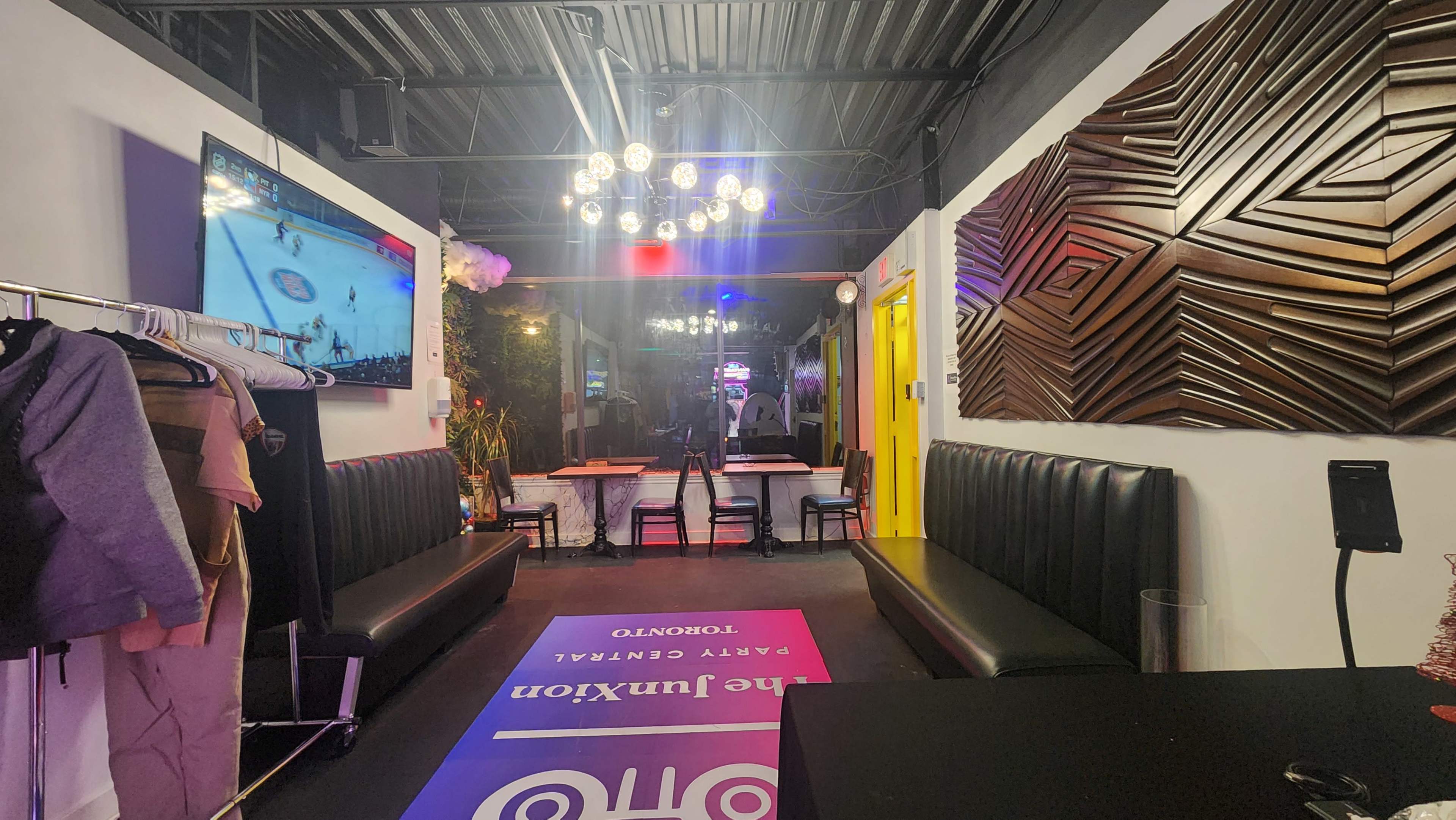 The image shows a modern interior of a lounge or entertainment space featuring black booths, colorful lighting, and a television displaying a sports game.