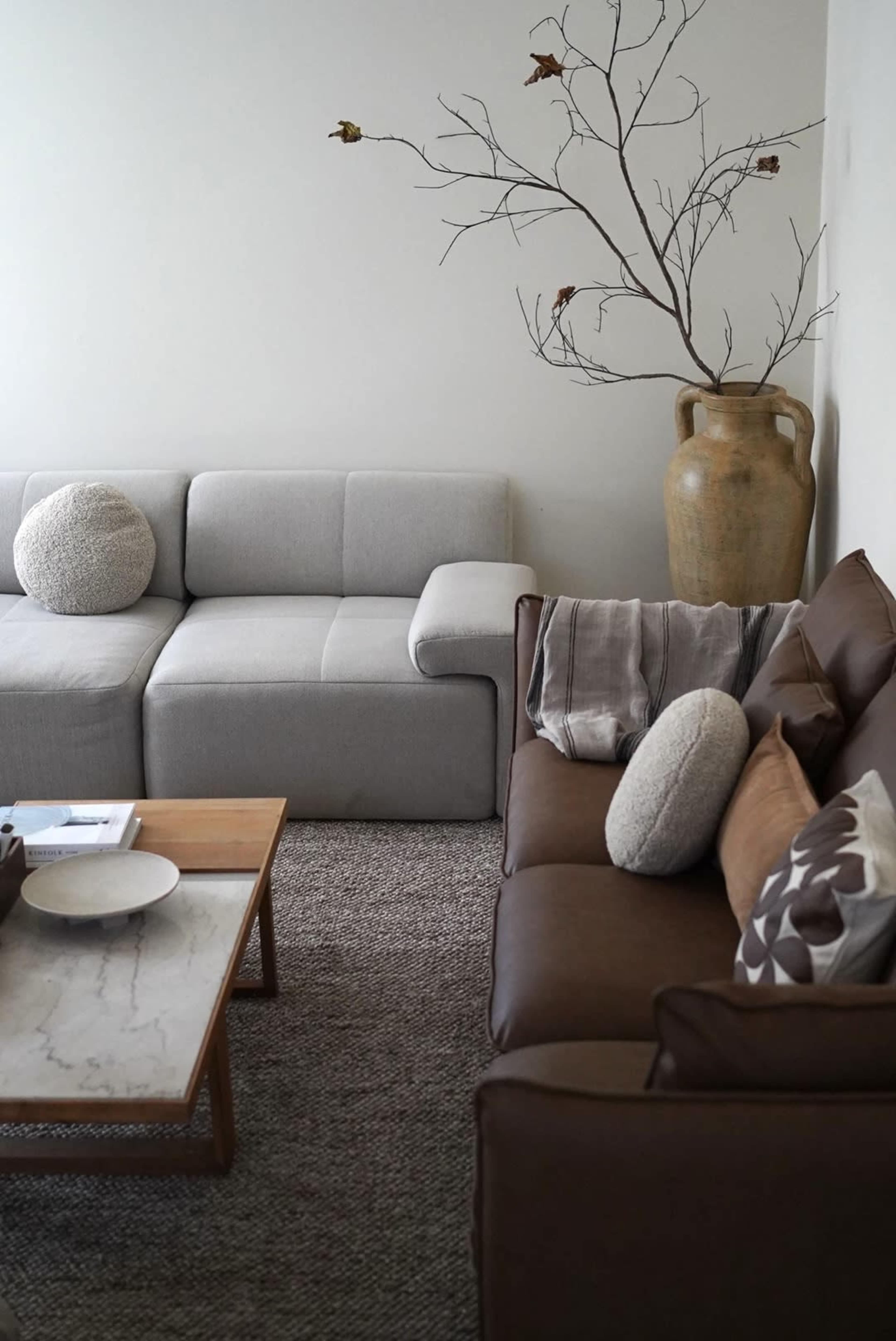 The image shows a living room with two sofas, one light gray and one brown, a wooden coffee table with a marble top, and a decorative vase beside a bare branch on the wall.