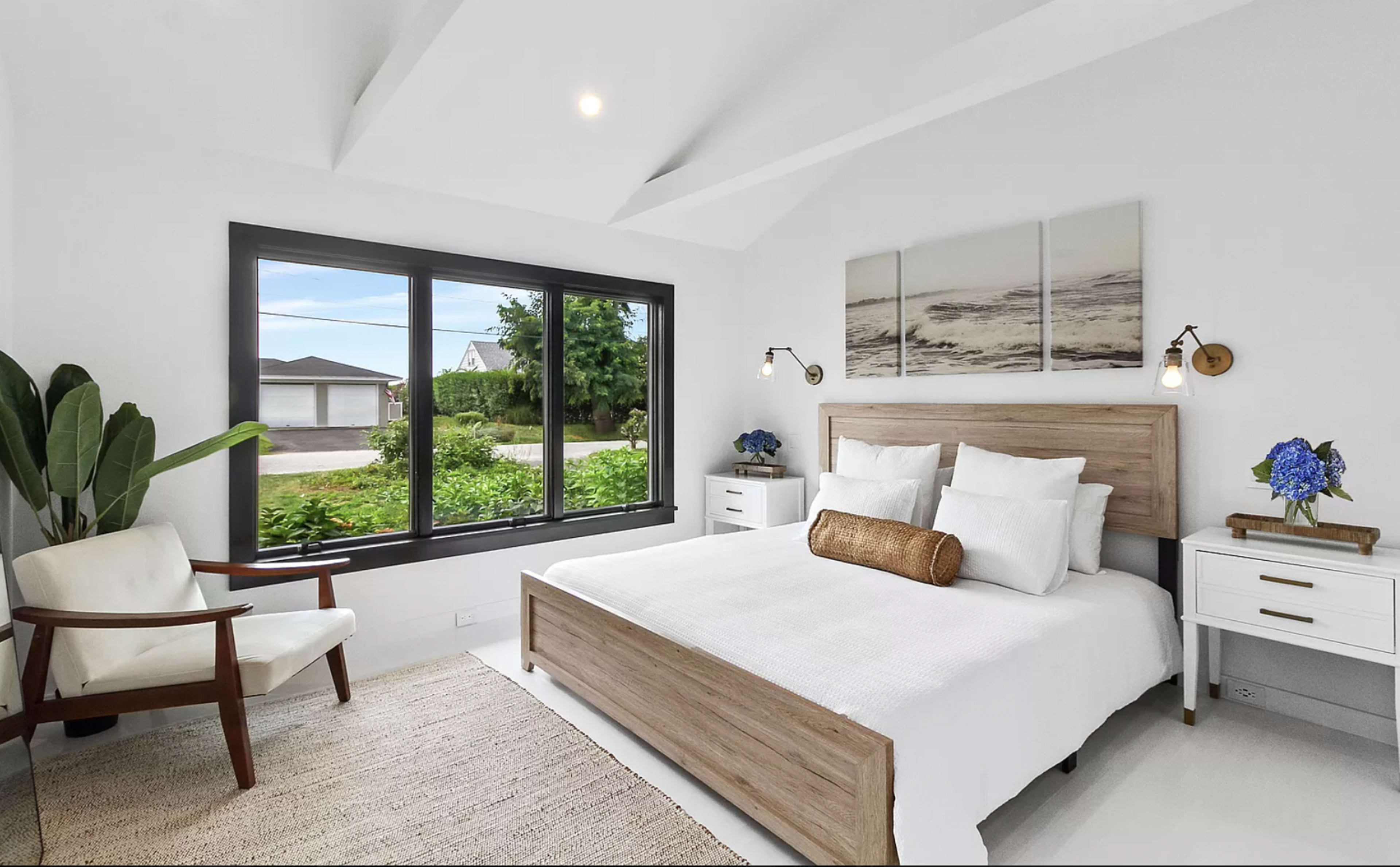 A bright, modern bedroom featuring a wooden bed frame, white bedding, and large windows that offer a view of greenery outside.