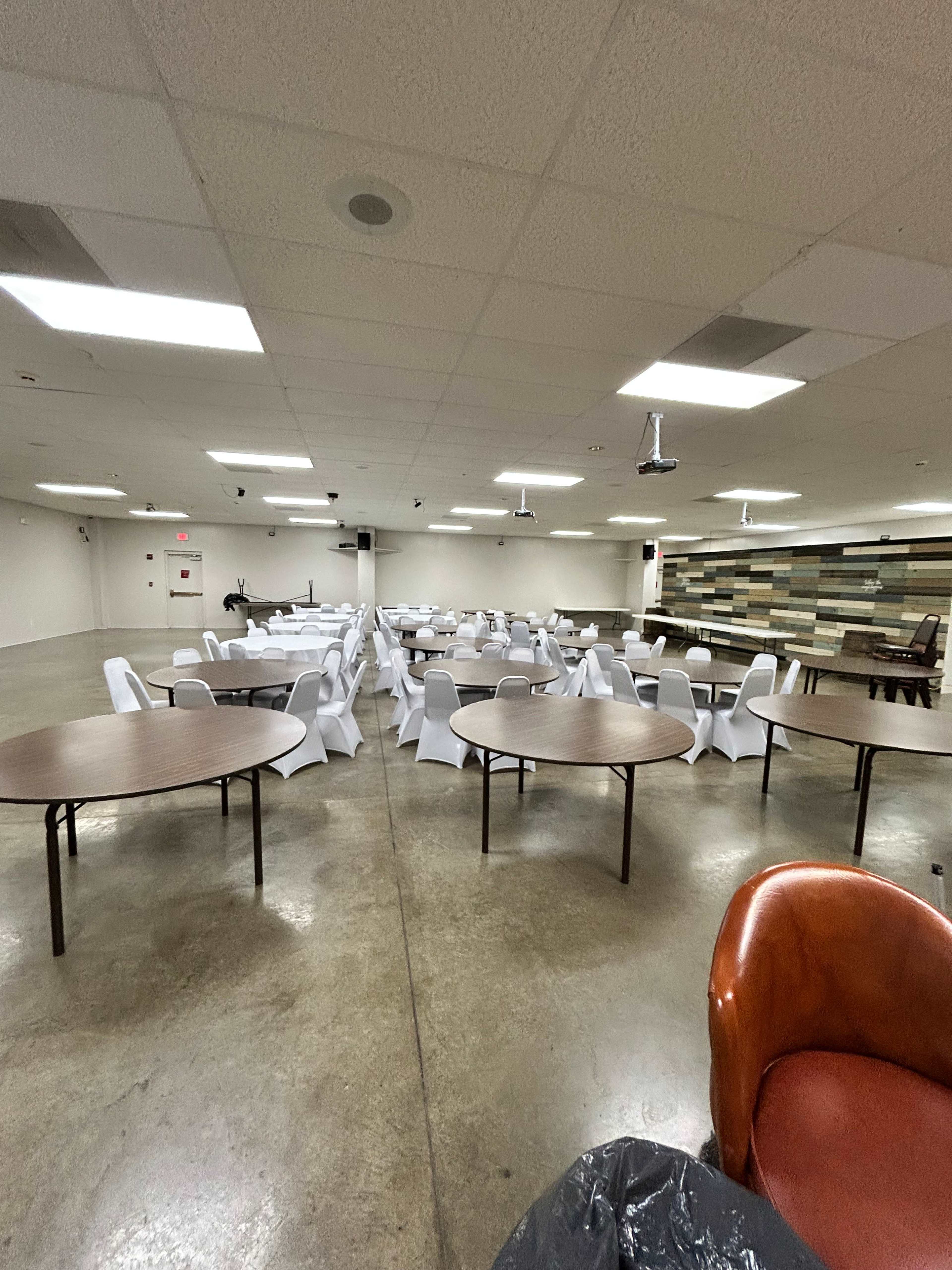The image shows a spacious indoor hall set up with several round tables and white chairs arranged for an event.