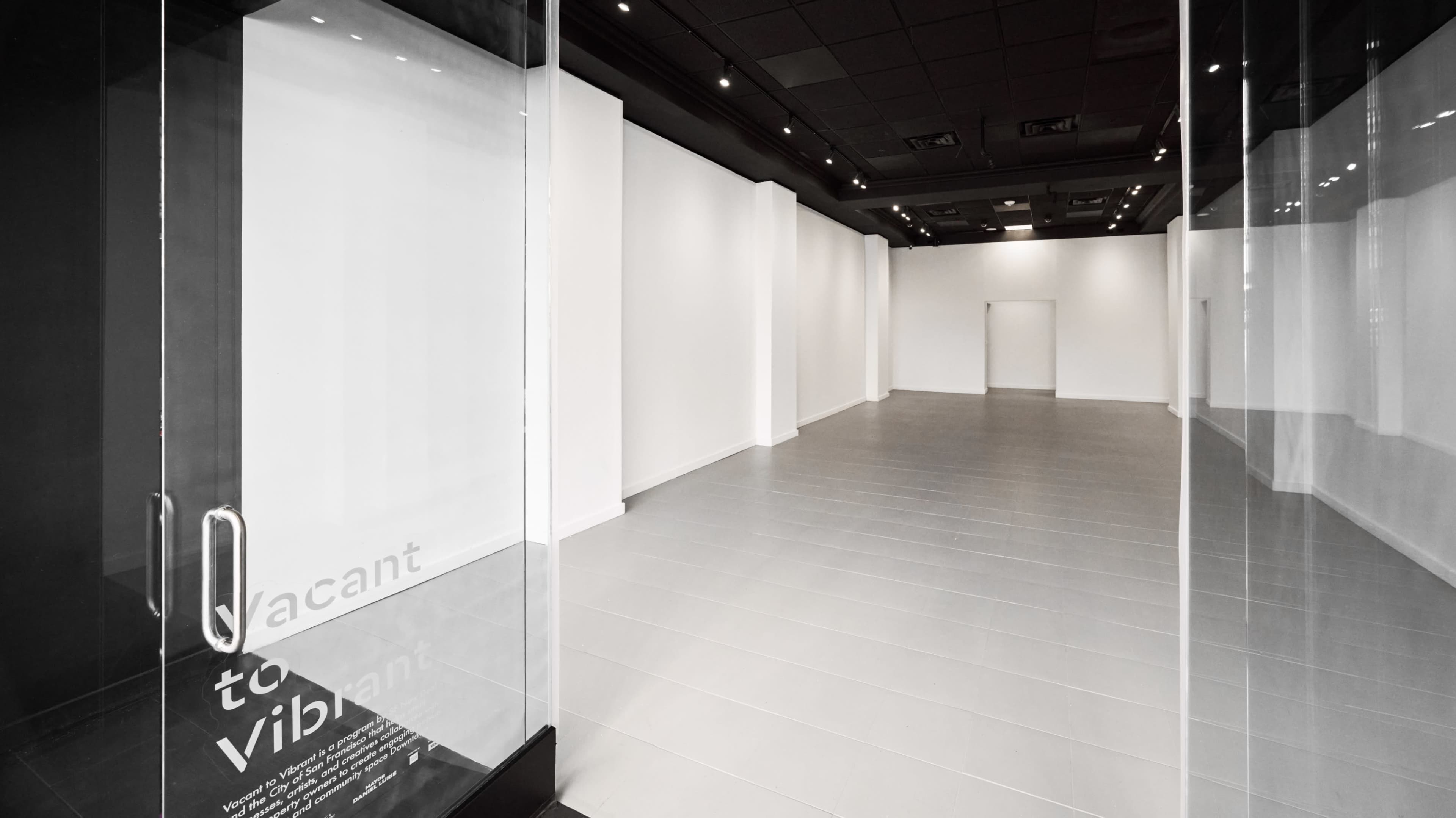 The image shows a vacant retail space with white walls and a gray tiled floor, featuring glass doors that provide an unobstructed view of the interior.