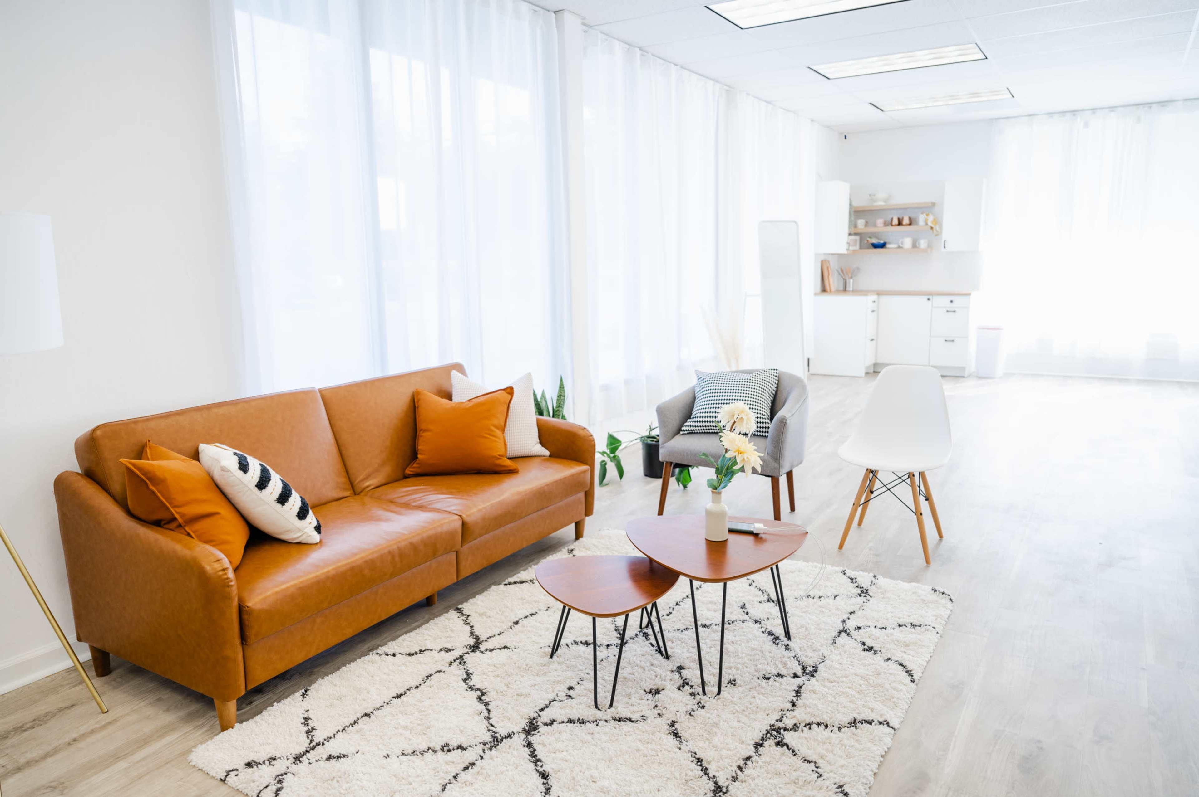 A bright and airy room features a brown leather sofa, two coffee tables, a gray armchair, and a modern kitchen area in the background.