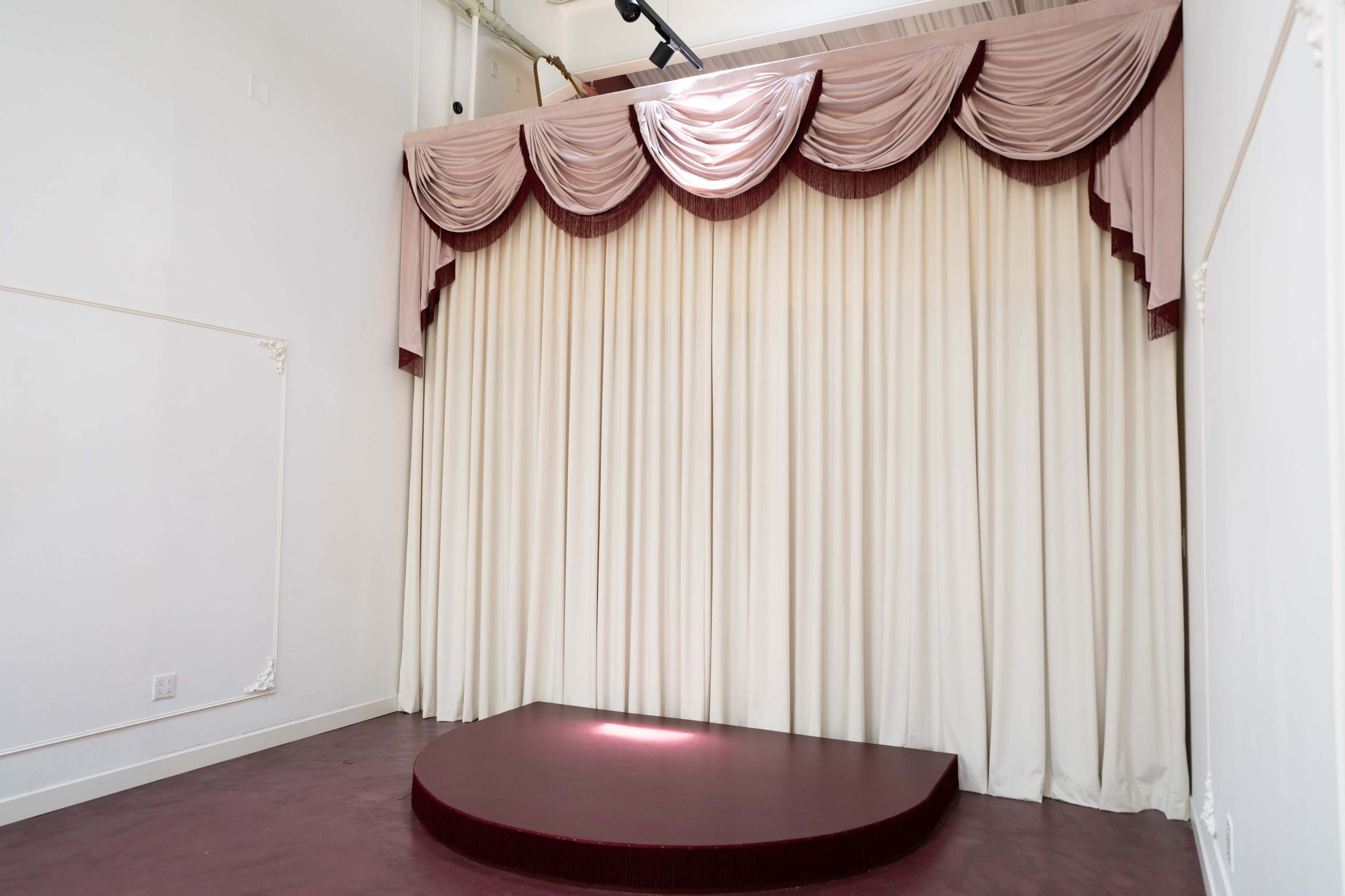 The image shows a small stage with a curved platform and a backdrop of draped curtains in a light color, accented with burgundy.