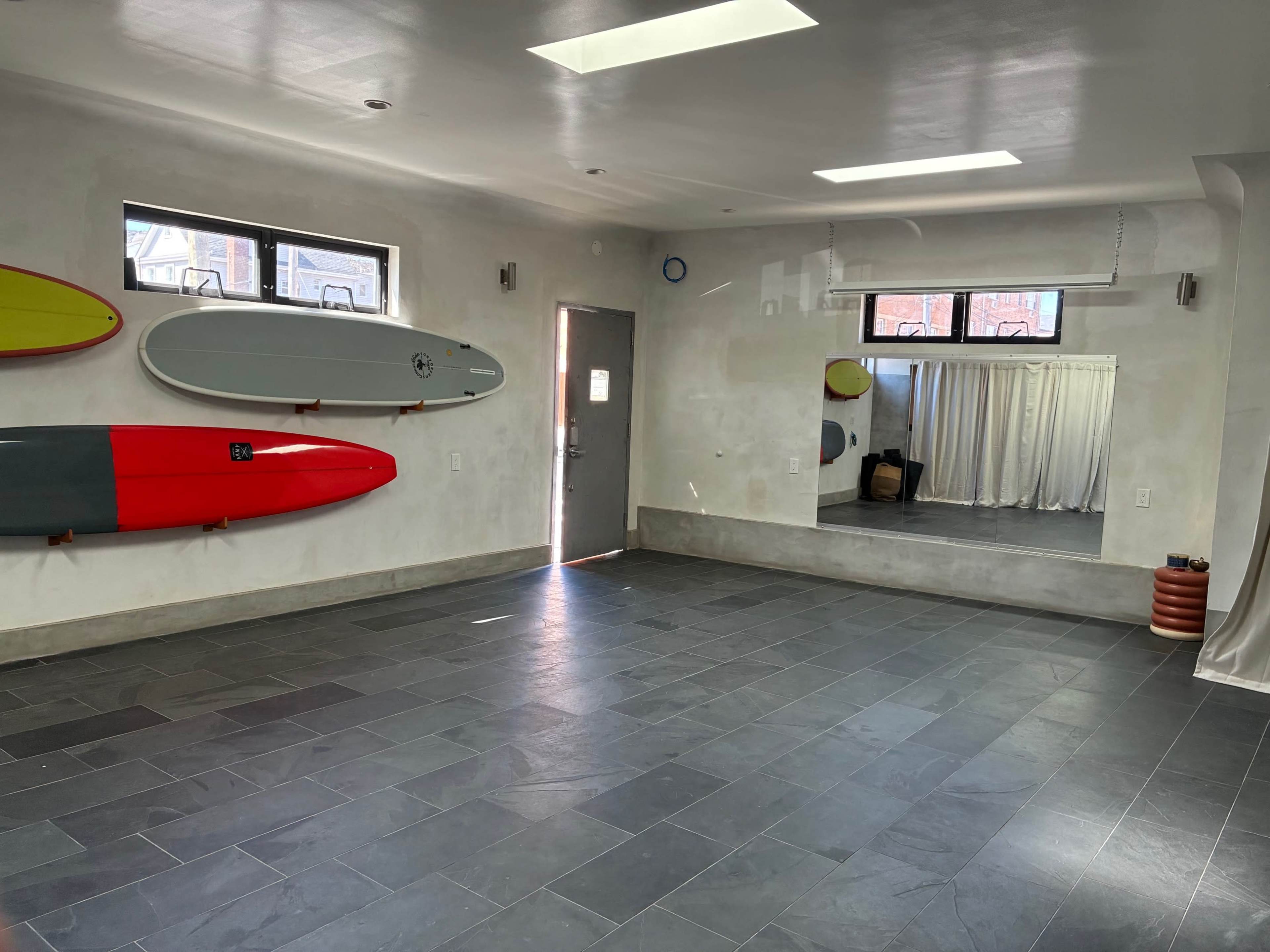 The room features a gray tiled floor, two windows, a mirror, and three surfboards mounted on the wall.