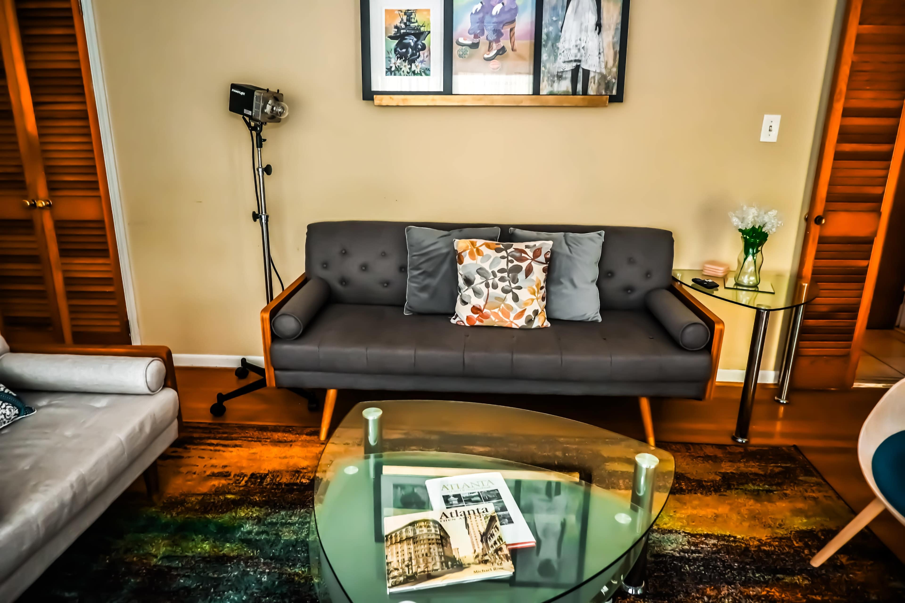 A modern living room features a gray couch with decorative pillows, a glass coffee table with magazines, and a light-colored area rug.