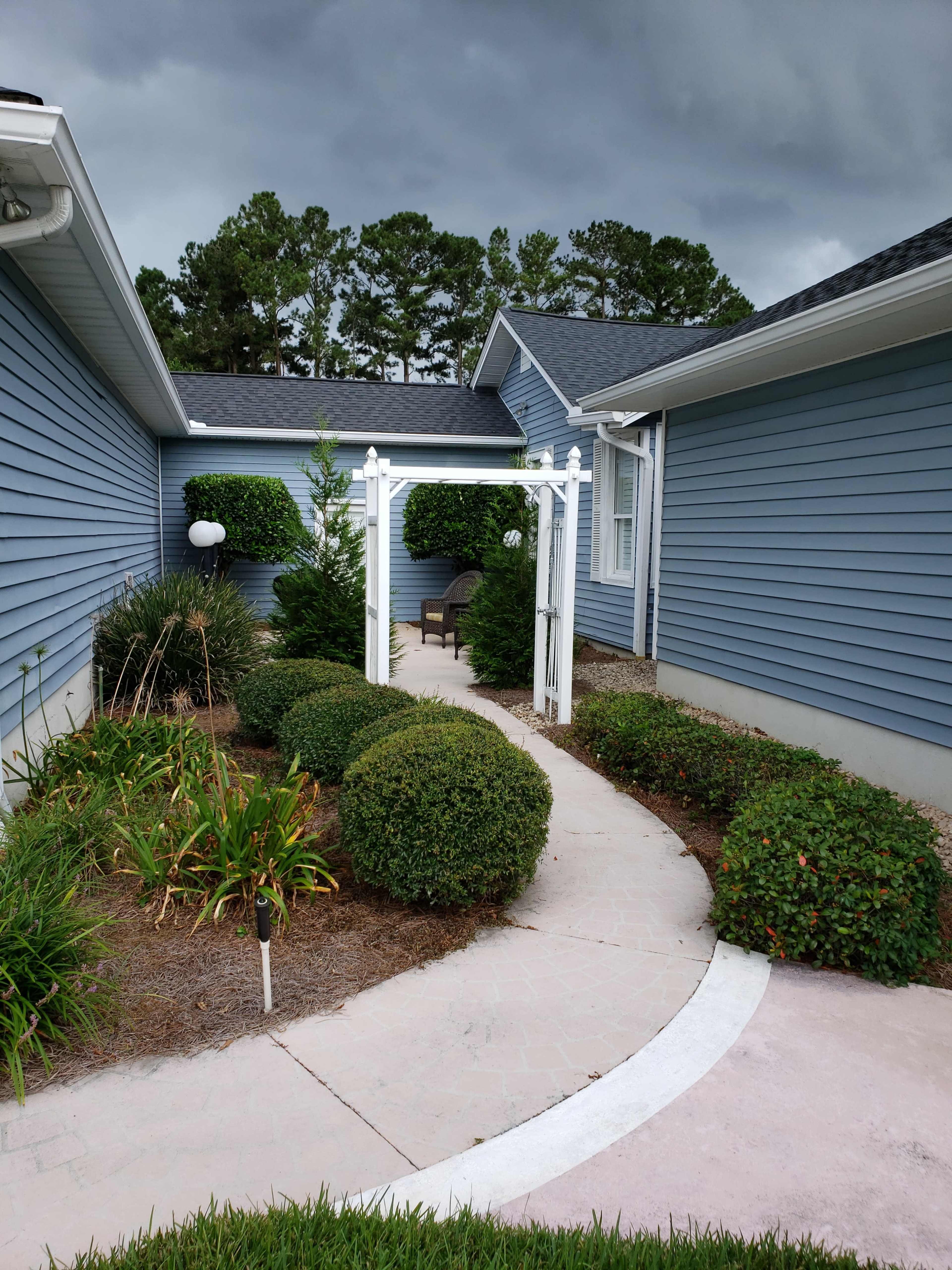 A curved pathway lined with neatly trimmed shrubs leads to a white arbor in a residential garden.