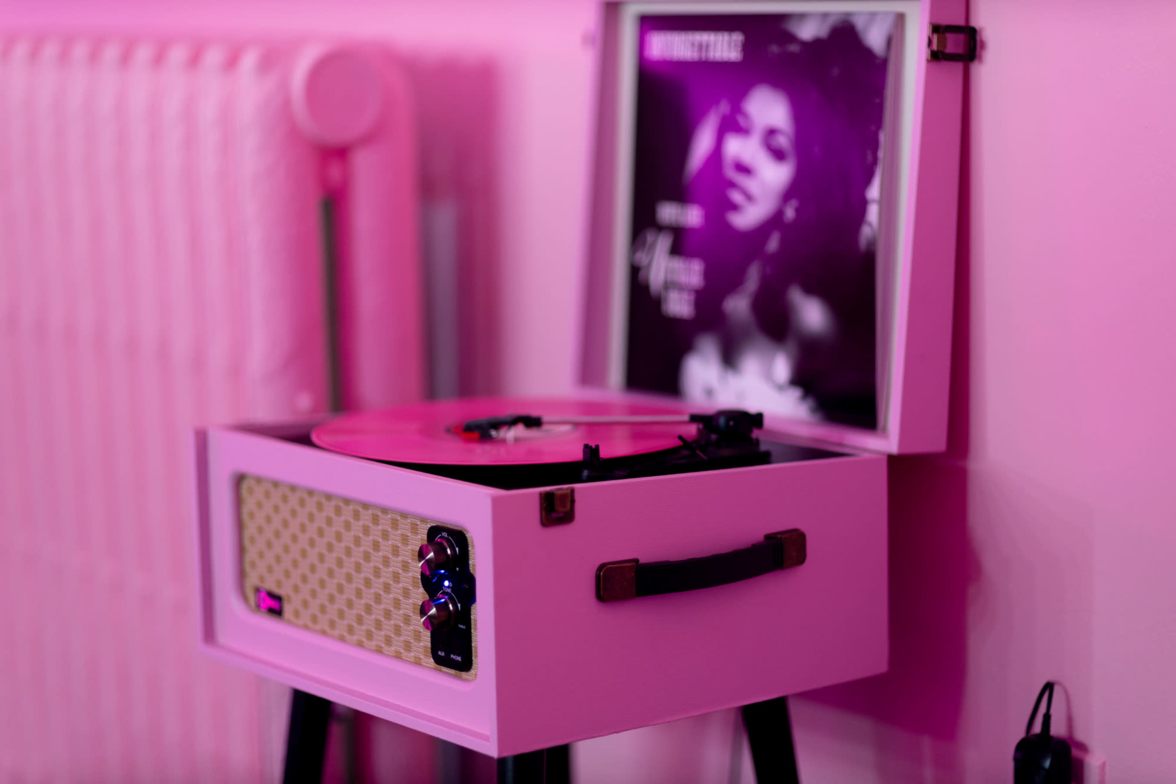 A pink turntable with a record playing and an album cover displayed stands on a small table against a pink wall.