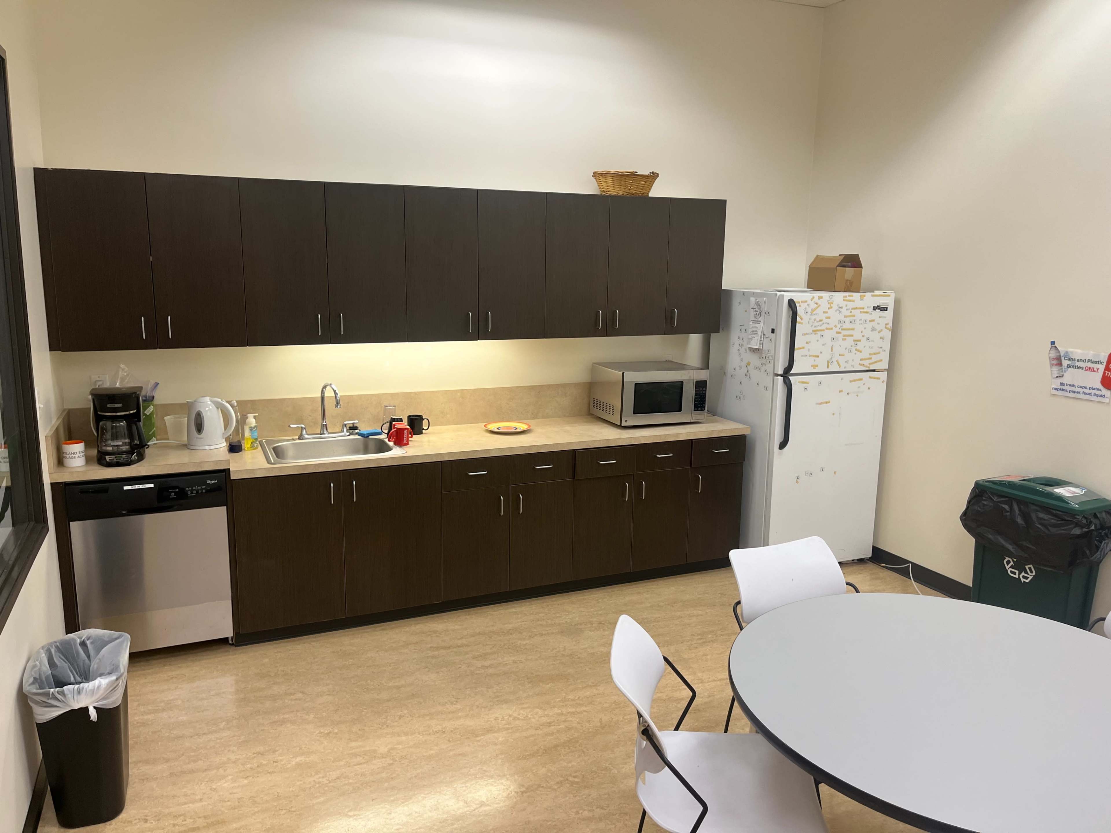 The image depicts a simple break room with a kitchen area that includes cabinets, a sink, a microwave, a refrigerator, and a small round table with chairs.