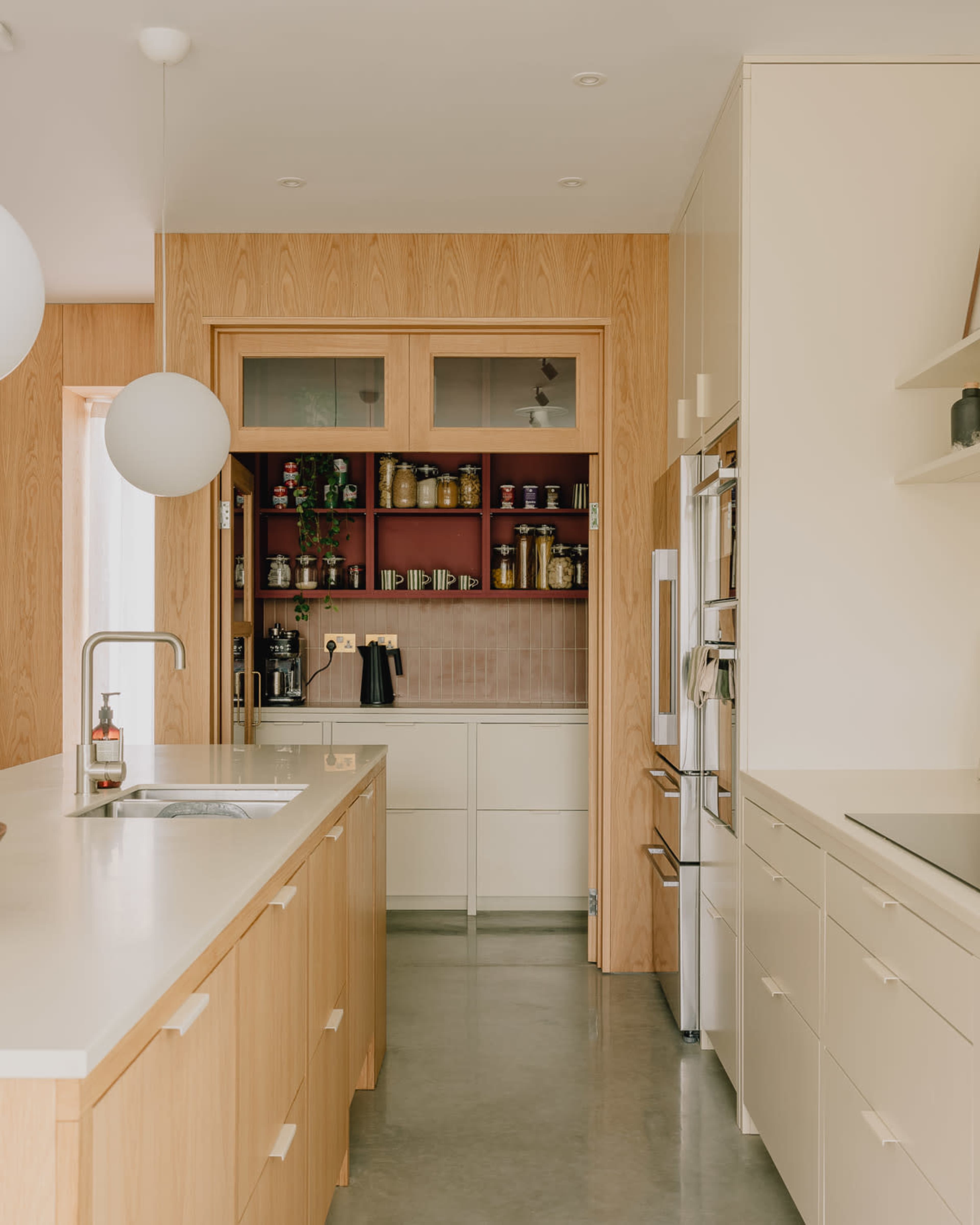The image shows a modern kitchen with wooden cabinets, a central island, and a pantry area featuring shelves with jars and kitchen appliances.
