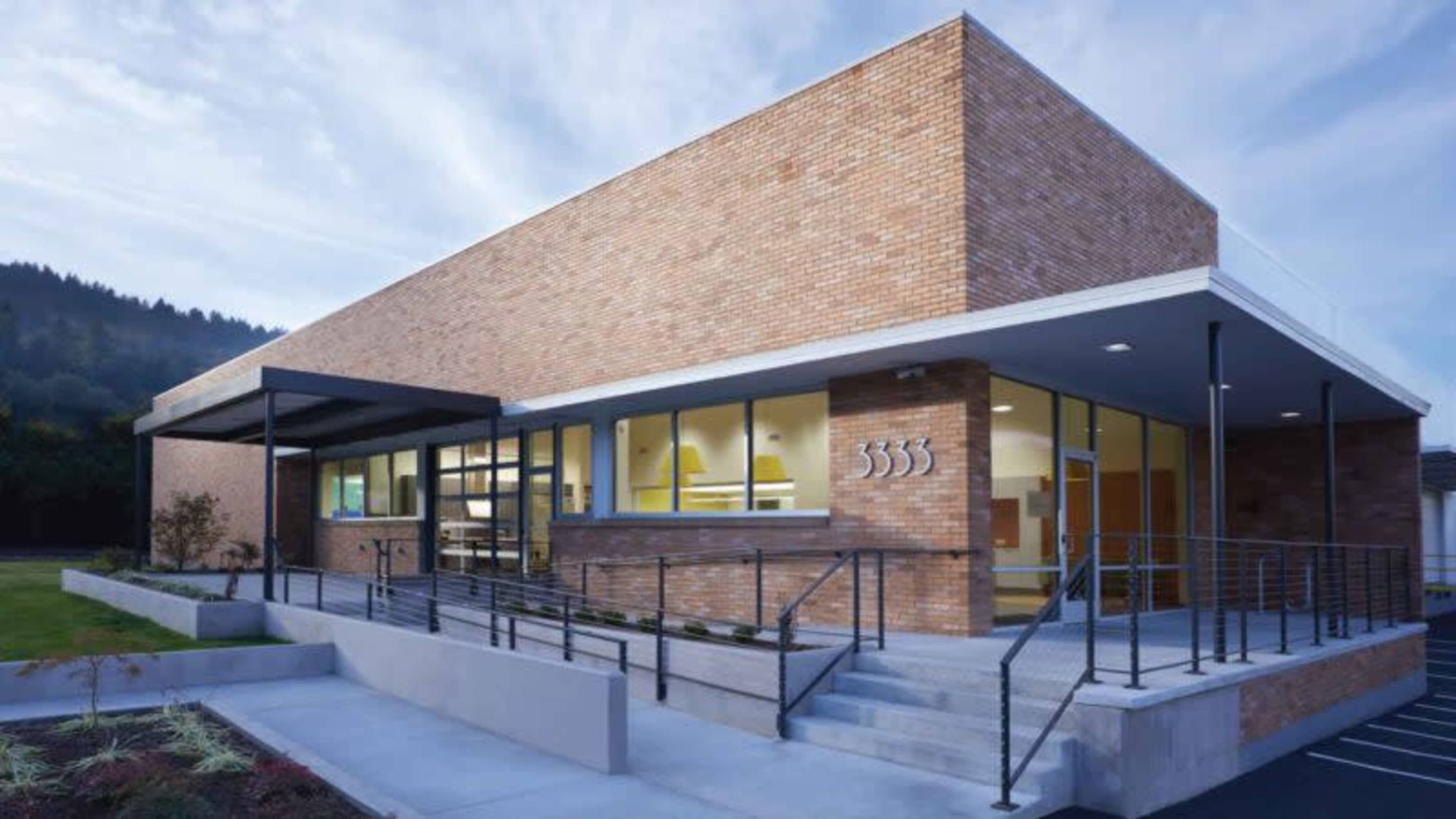A modern, brick building with large windows and a staircase leading to the entrance, set against a backdrop of greenery and a cloudy sky.