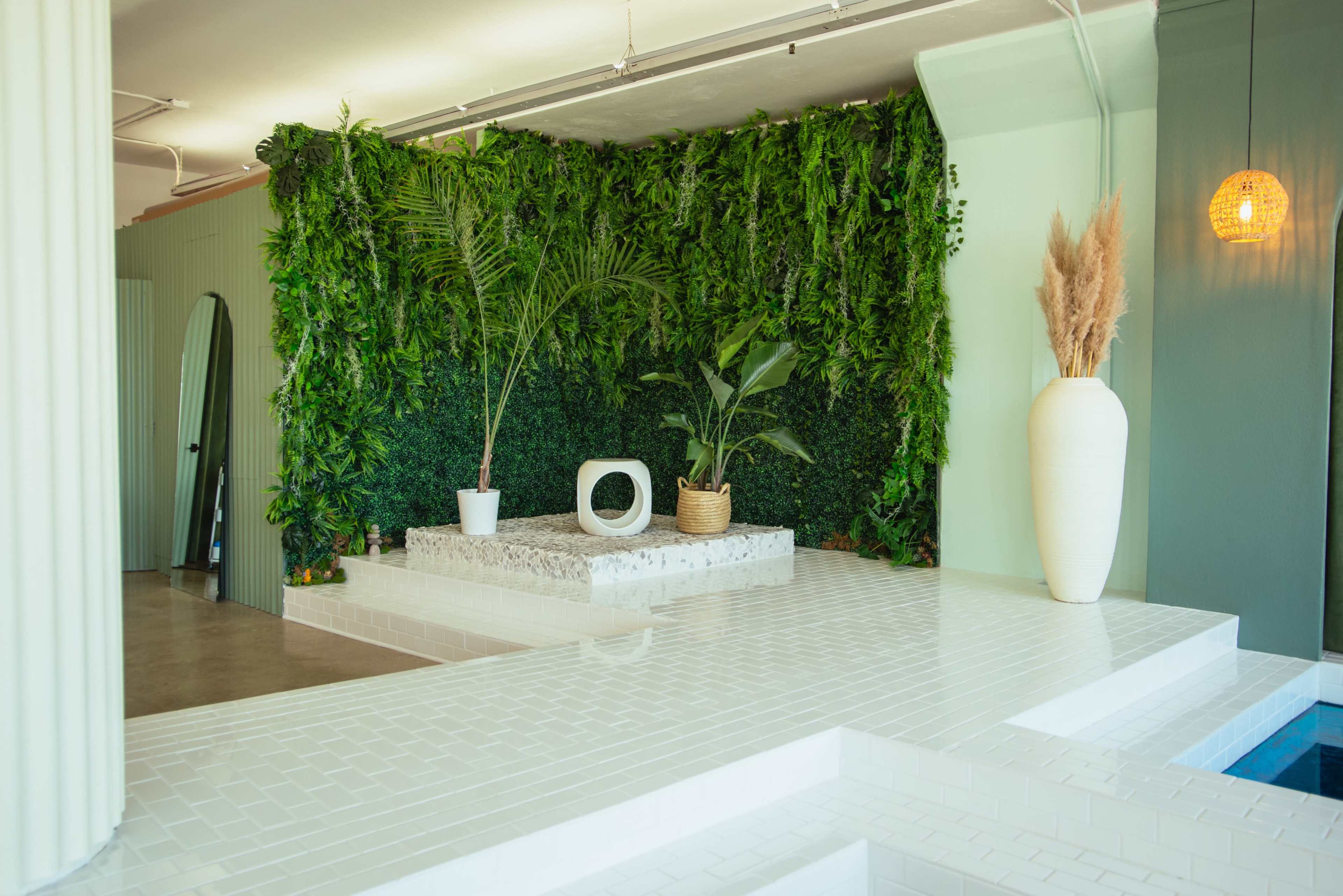 A modern interior space featuring a white tiled platform with plants against a green wall, accented by a large white vase and a curved mirror.