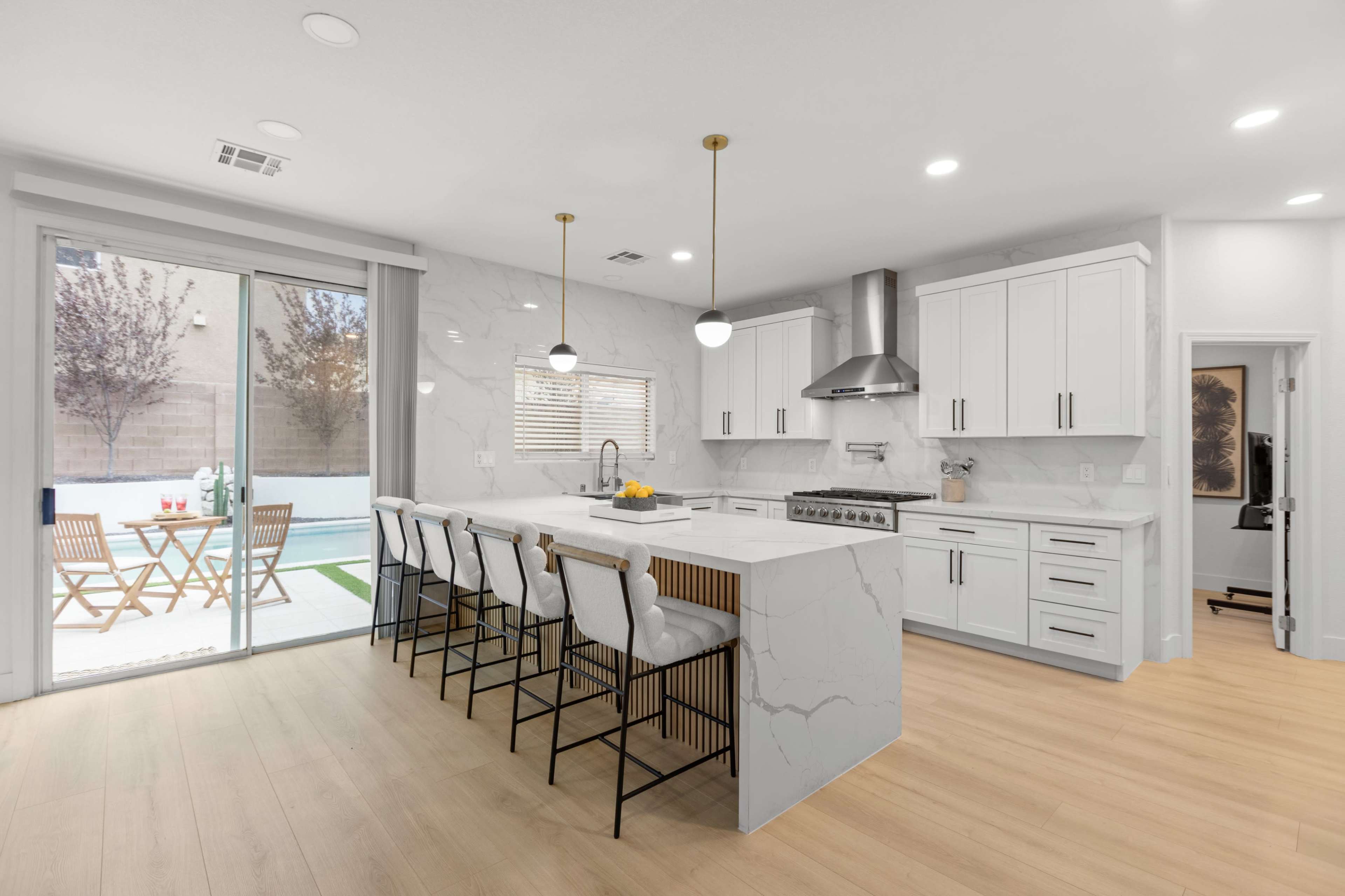 The image shows a modern kitchen featuring a large marble island with bar stools, white cabinetry, and a sliding glass door that opens to an outdoor seating area.