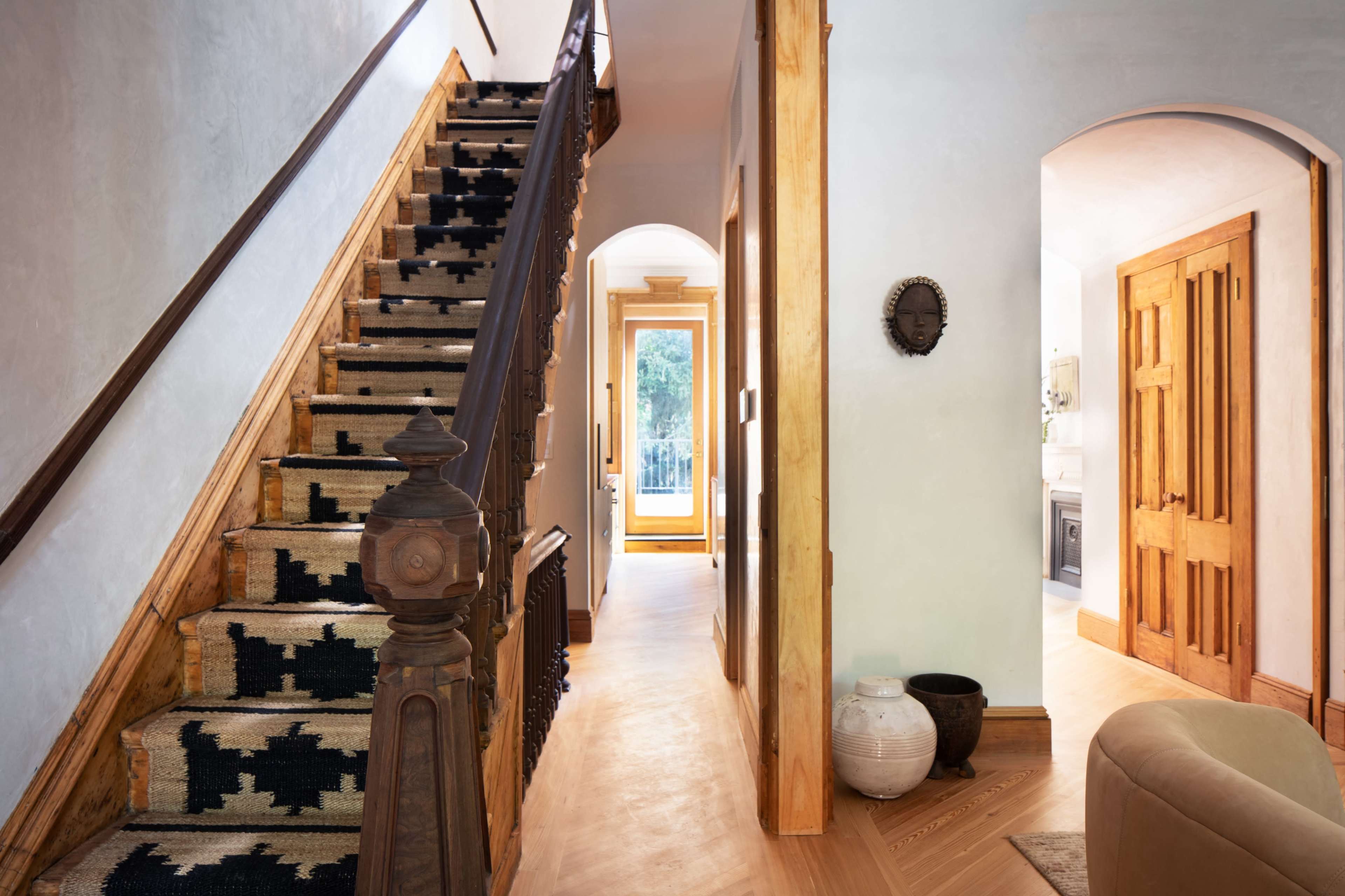 The image shows a staircase with a patterned carpet leading to an upper level, alongside a hallway that connects to a bright room with a door open to an outdoor view.