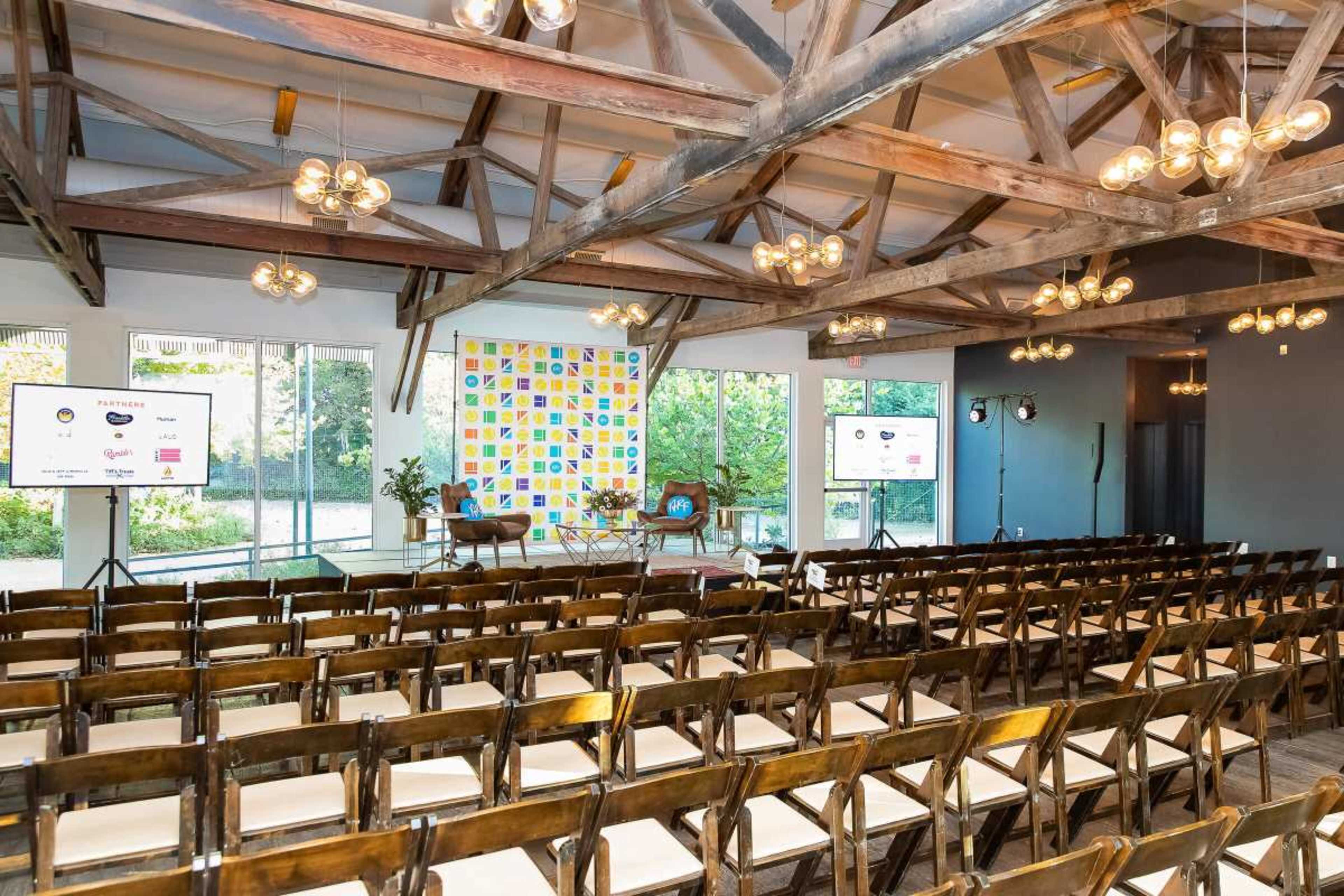 A modern event space features rows of empty chairs facing a stage with two armchairs, flanked by large screens and colorful patterned decor.