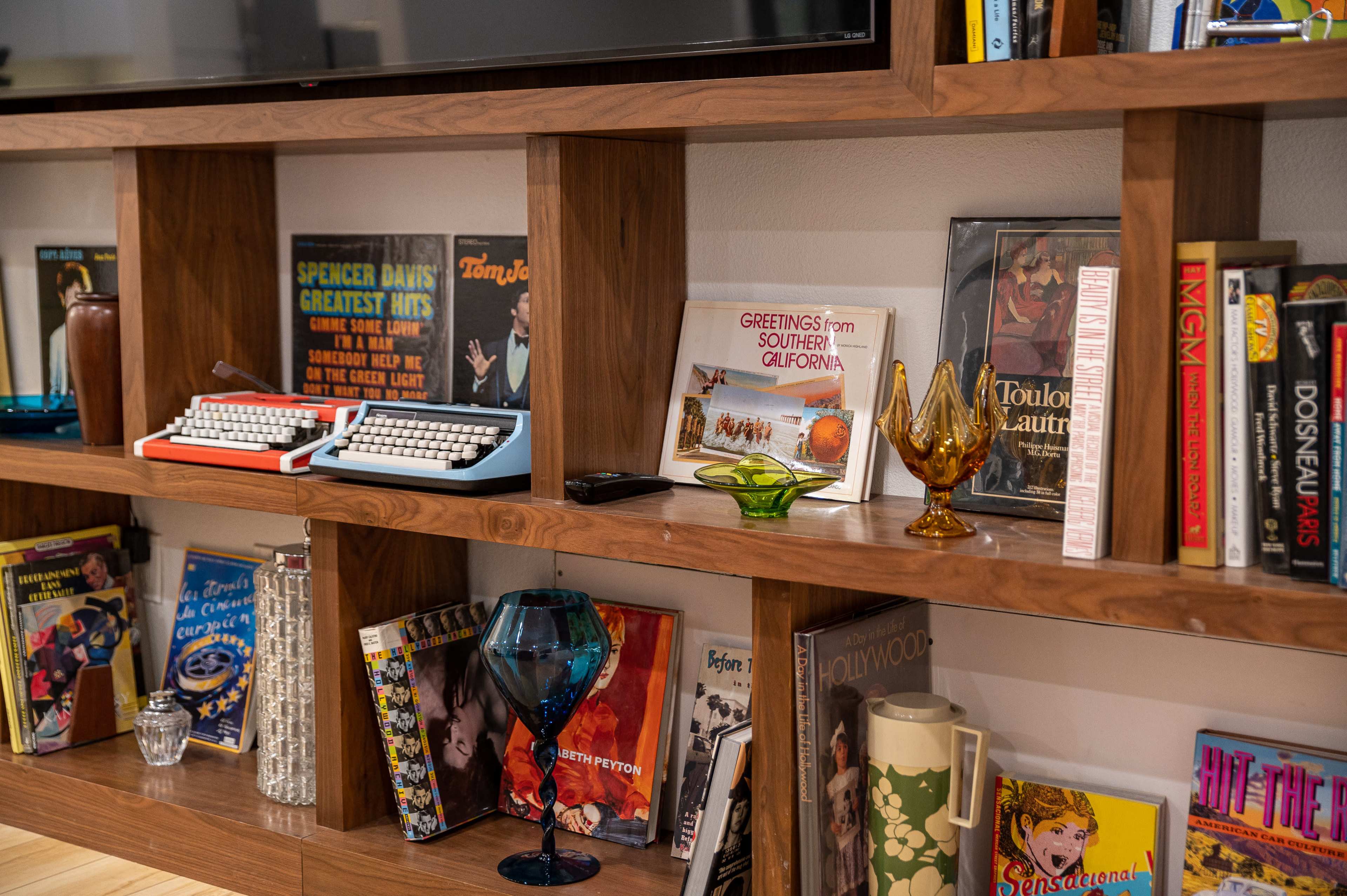 A wooden shelf displays an assortment of vintage items, including a typewriter, books, glassware, and postcards.