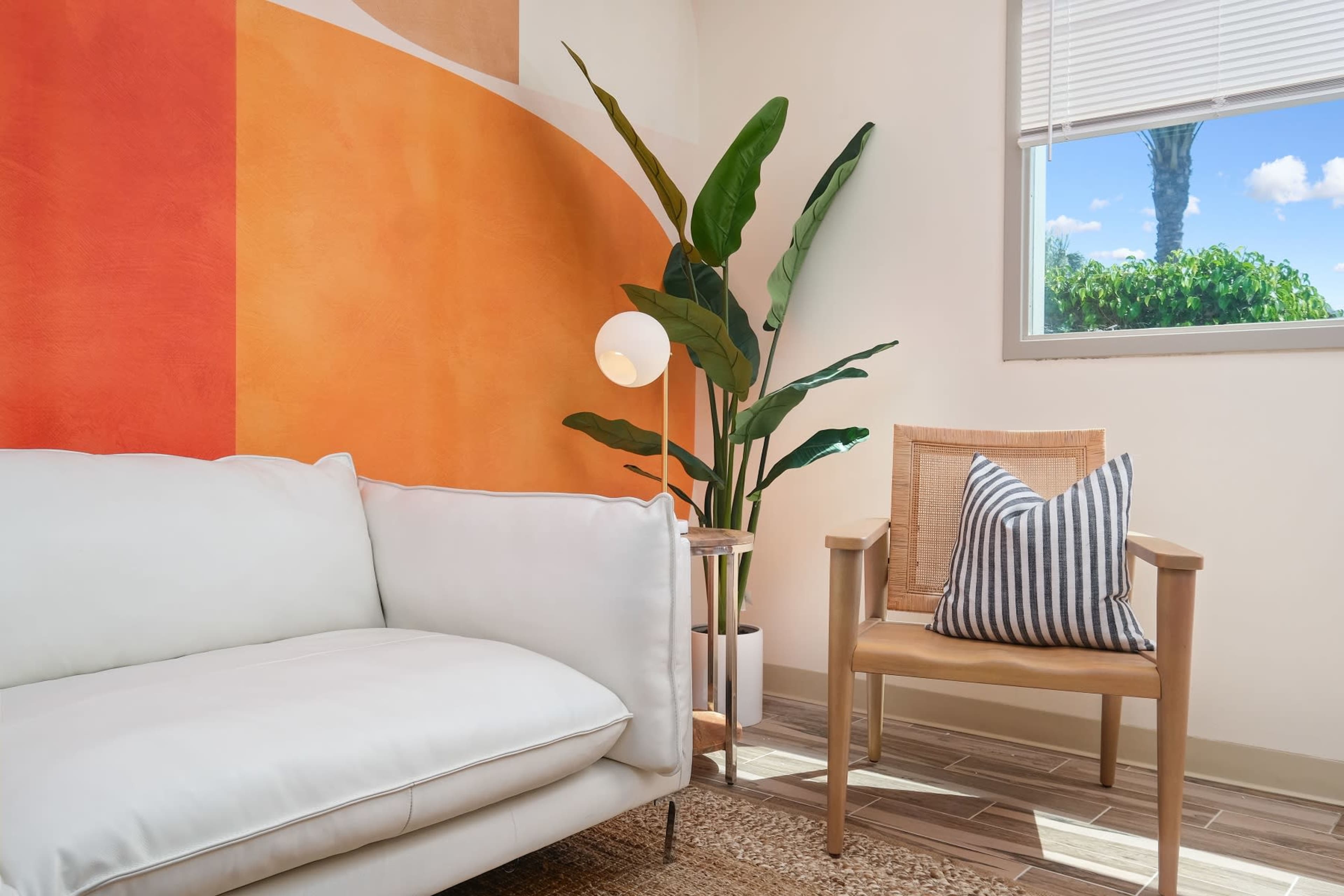 A modern living area features a white couch and a wooden chair beside a plant, with an orange wall mural and a window showing blue skies.